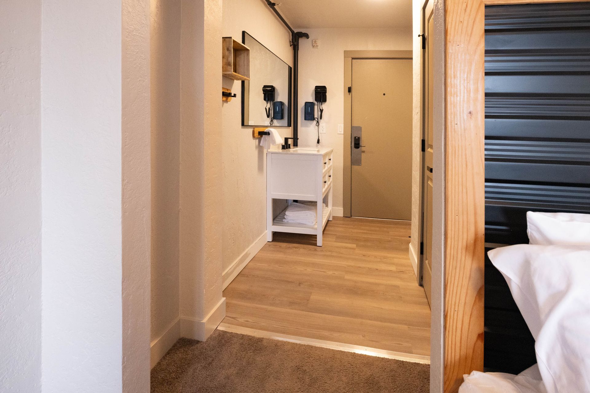 Hotel room hallway with light wood flooring, a small white desk, and a bed visible on the right.