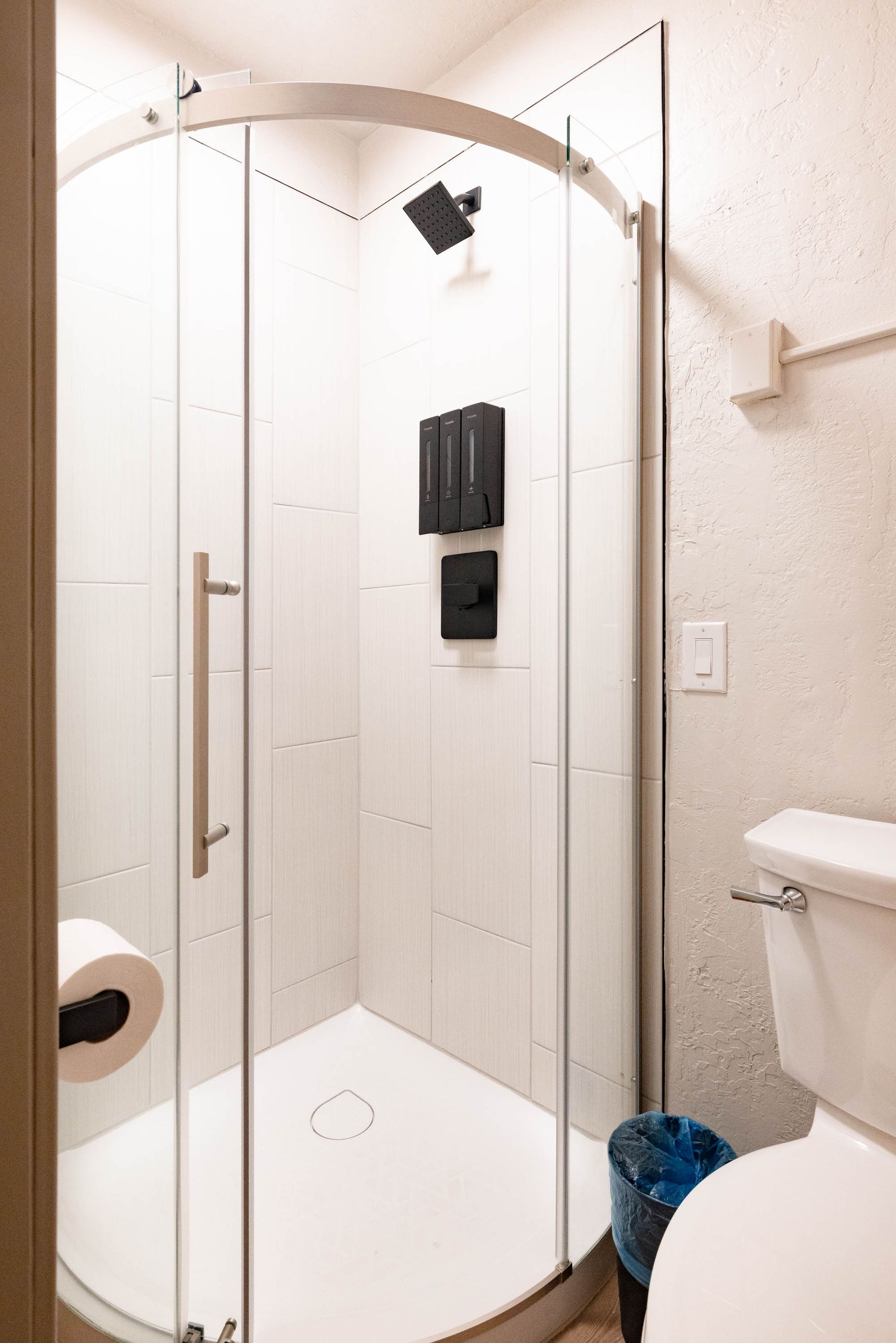 Small bathroom shower with clear glass door, white tile, overhead shower, and toilet visible beside it