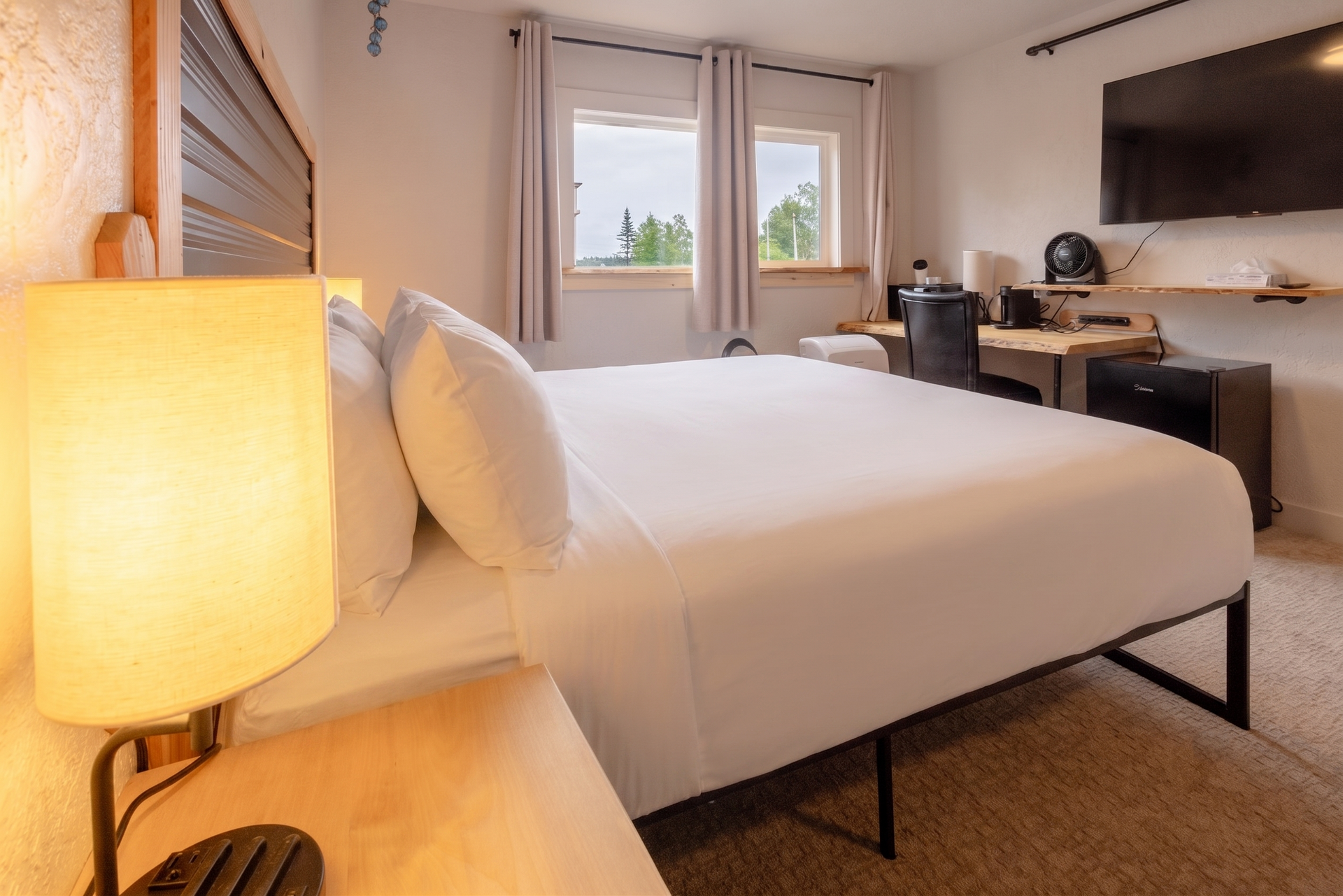 Modern bedroom with a neatly made white bed, bedside lamp, desk, and window view