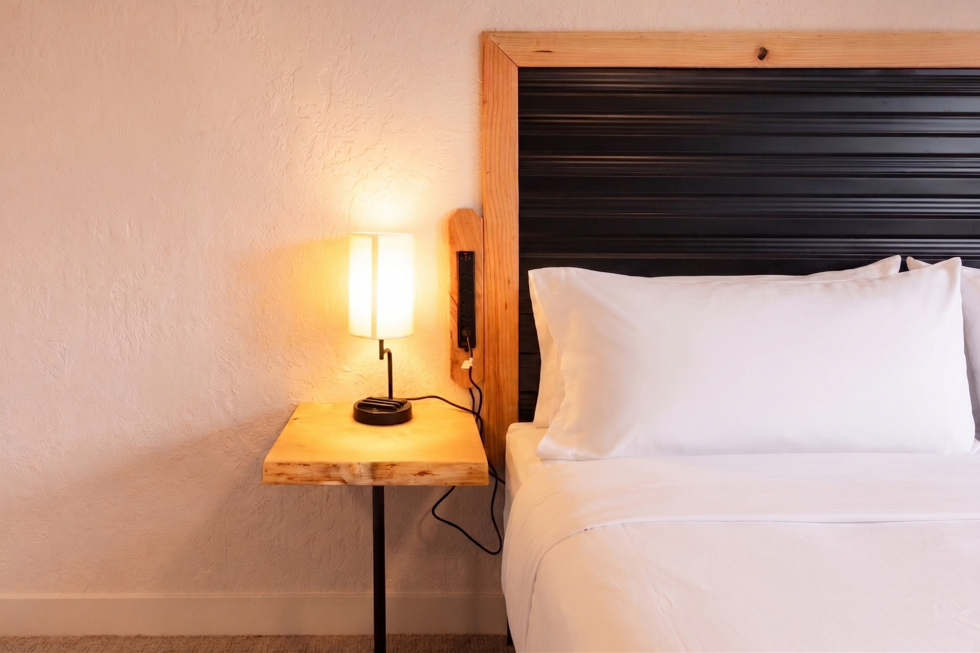 Bed with white pillows beside a lit lamp on a wooden nightstand in a hotel room