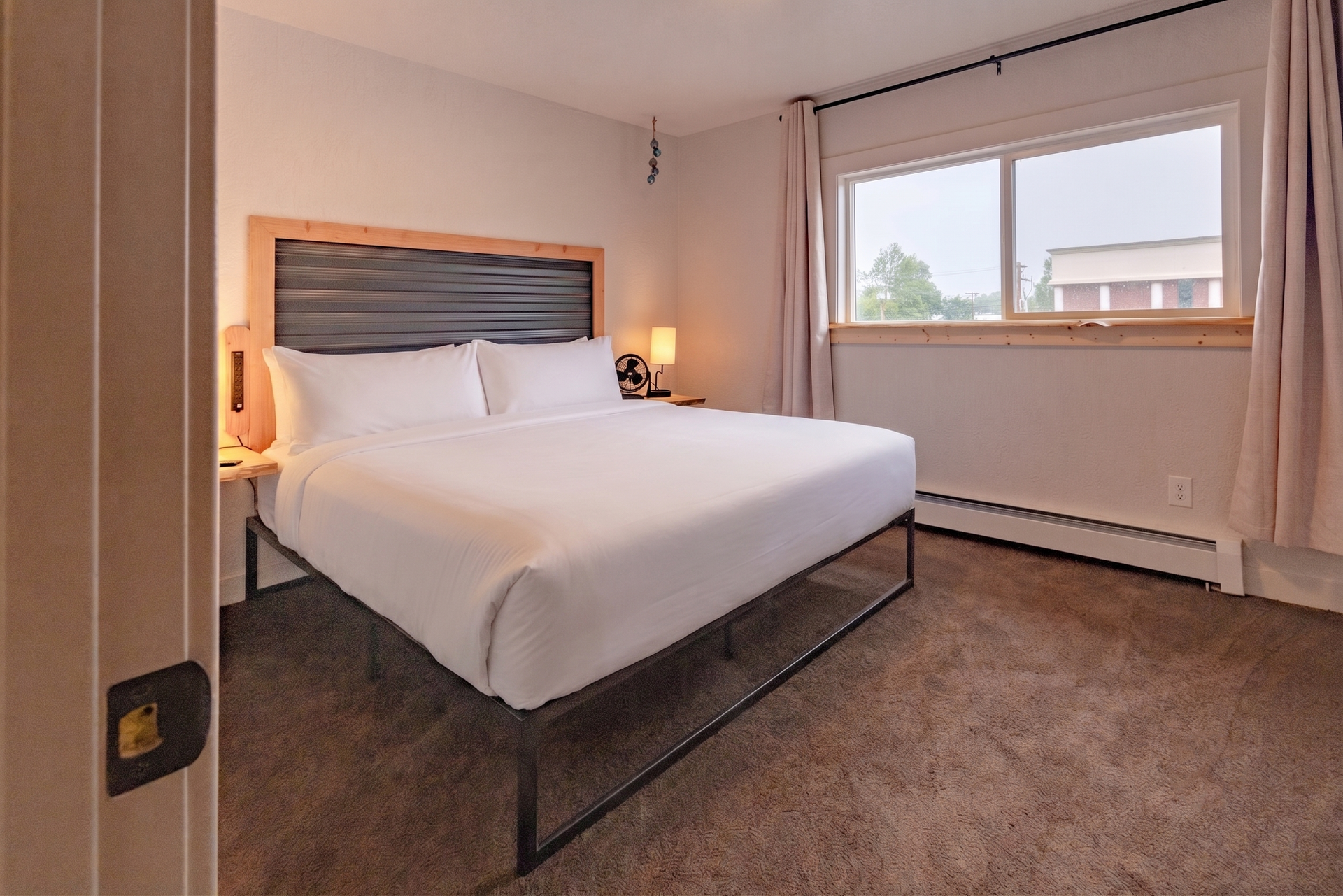Small bedroom with a made bed, nightstands, and a large window showing an apartment building.