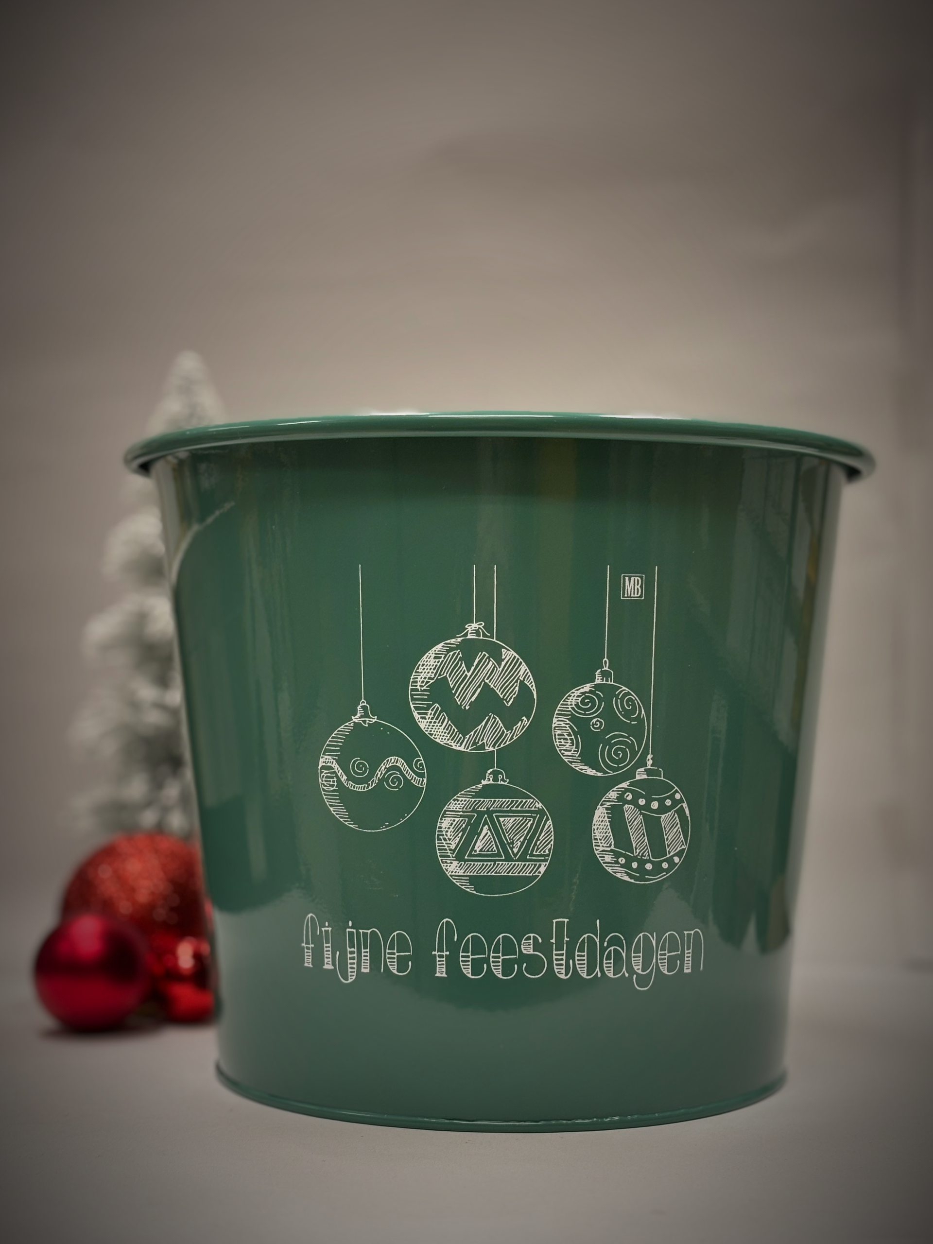 Groene emmer met witte kerstversieringen en tekst. Rode versieringen en een kleine boom op de achtergrond.