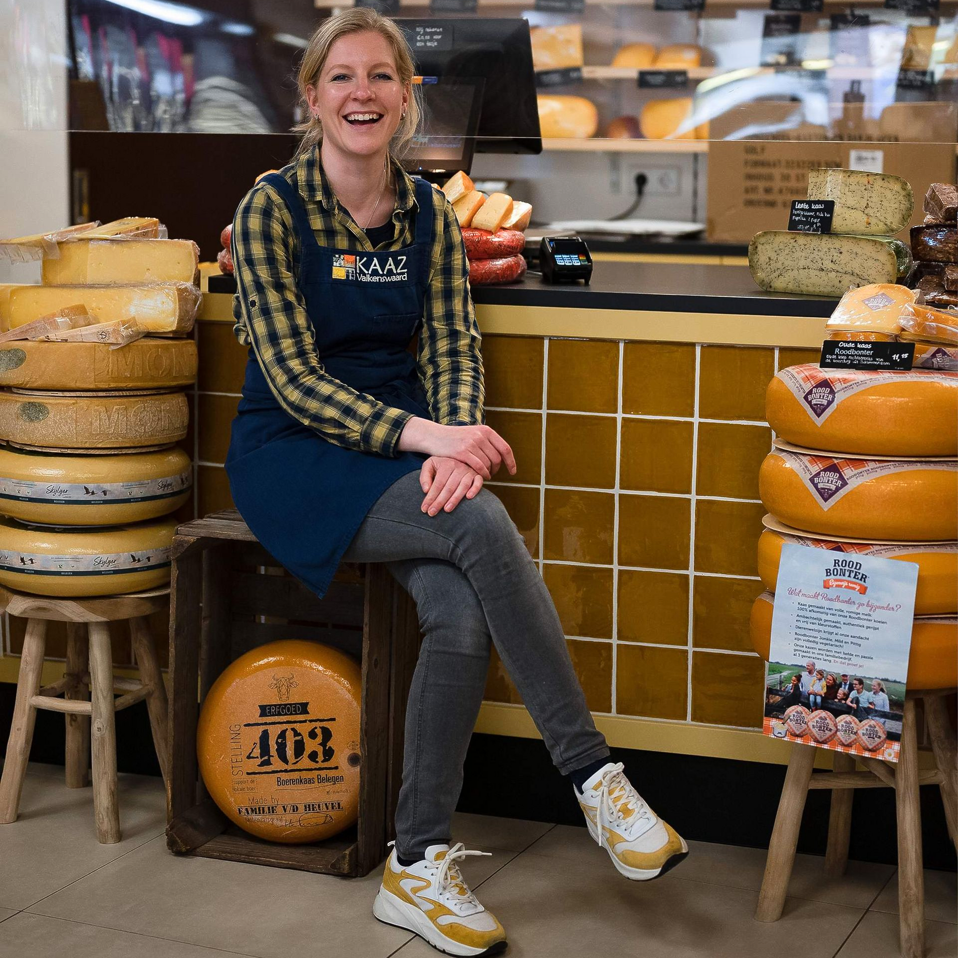 Lachende vrouw zit in een kaaswinkel, draagt ​​een schort en is omringd door kazen.