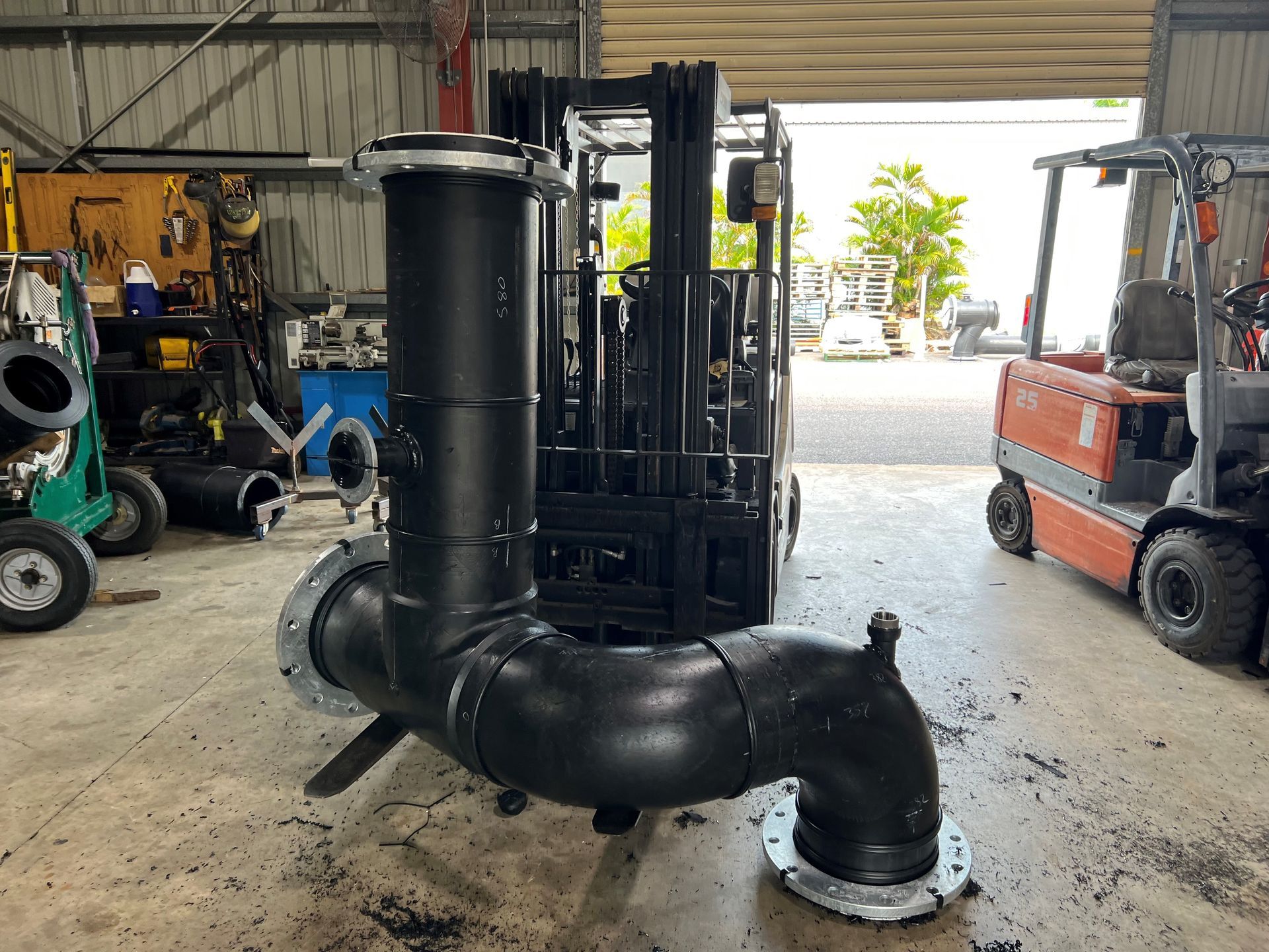 Black industrial pipe assembly with large curved sections in a warehouse, flanked by forklifts. — Advanced Poly in Darwin, NT
