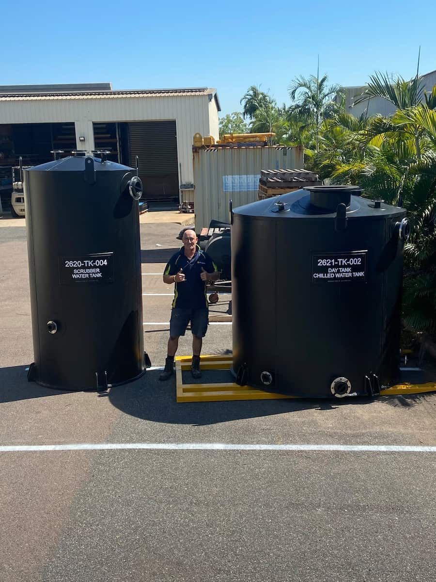 A person stands between two large, black industrial tanks in an outdoor lot, giving a thumbs up. — Advanced Poly in Yarrawonga, NT