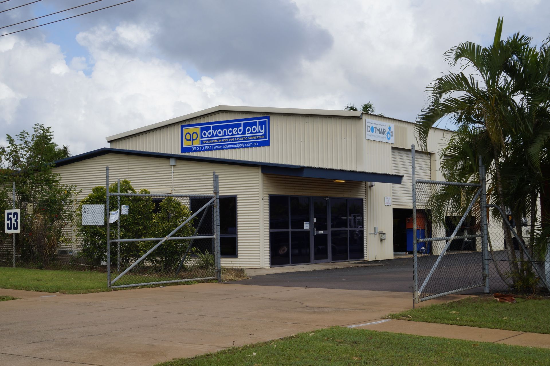 Small commercial building with a blue sign and chain-link fence, seen from the street under cloudy skies — Advanced Poly in Darwin, NT