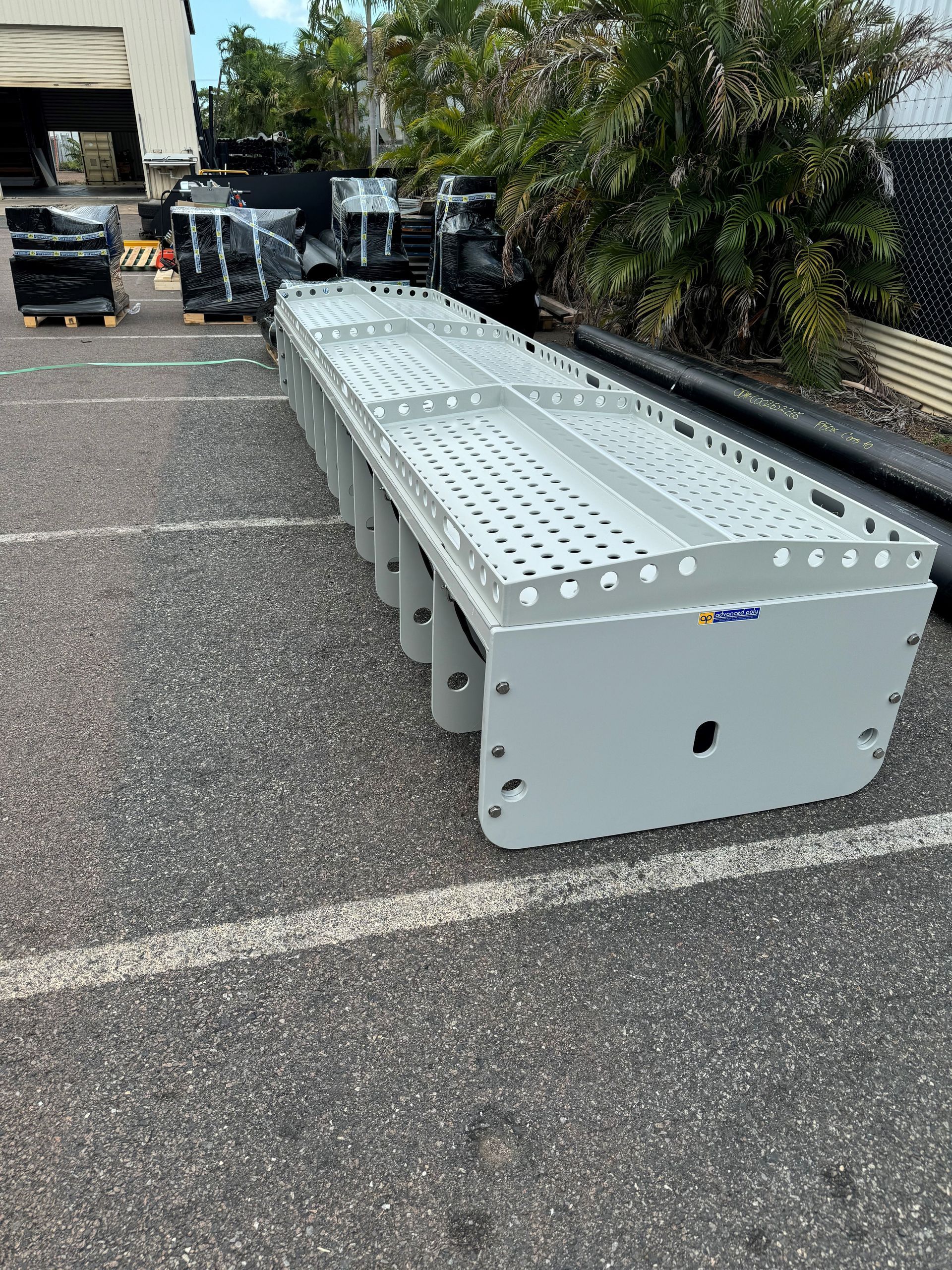Long metal equipment rack on asphalt in a parking lot, with parked vehicles and greenery nearby — Advanced Poly in Darwin, NT