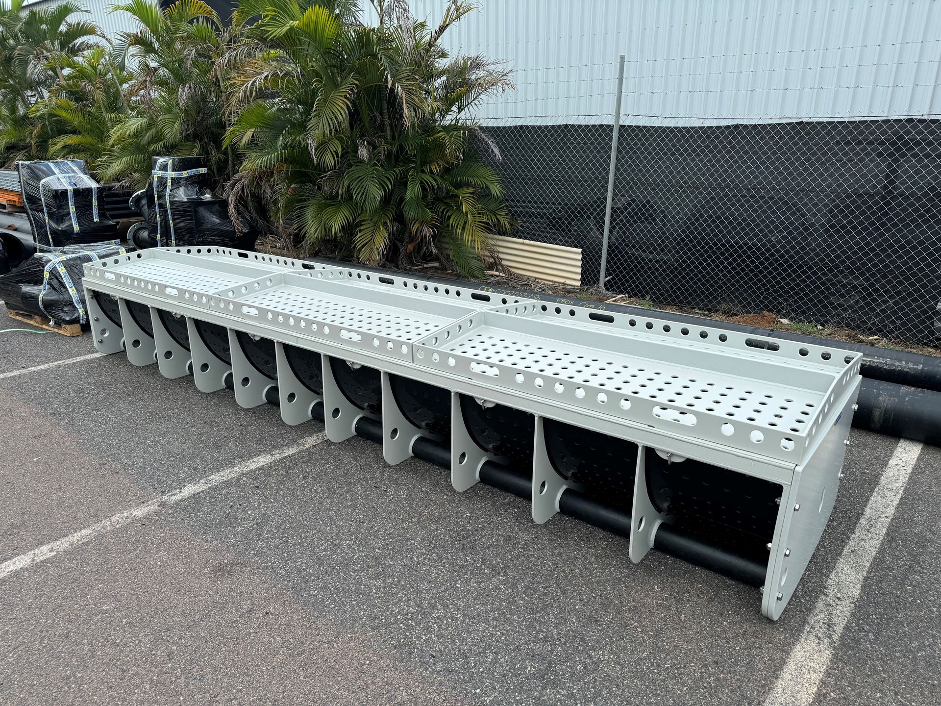 Long metal industrial ramp with wheel-like supports, leaning outdoors beside a fence and palm trees — Advanced Poly in Darwin, NT