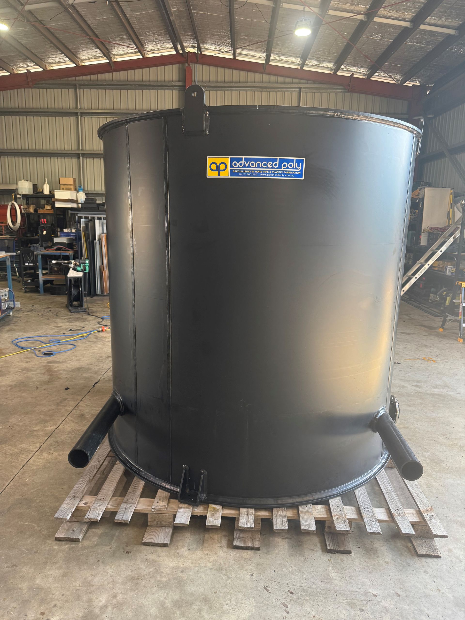 Large black cylindrical metal tank in a workshop, sitting on wooden pallets under a corrugated roof. — Advanced Poly in Darwin, NT — Advanced Poly in Darwin, NT — Advanced Poly in Darwin, NT — Advanced Poly in Darwin, NT — Advanced Poly in Darwin, NT 