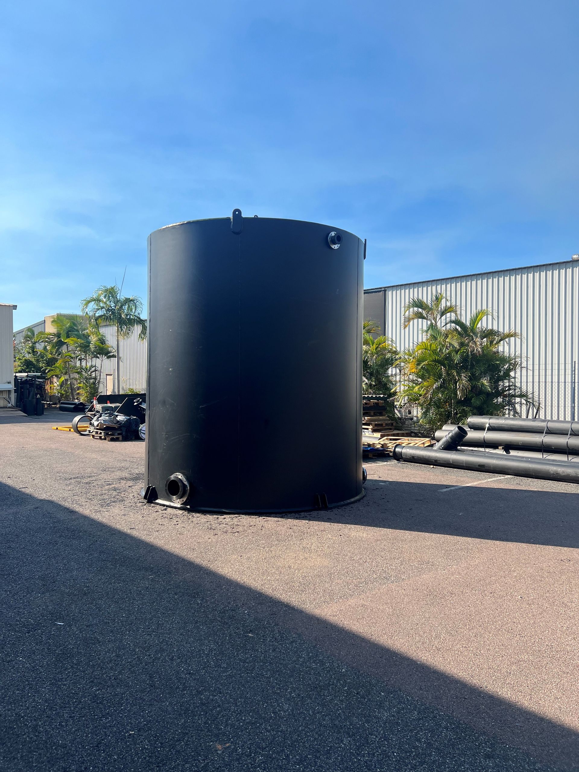 Large black cylindrical tank on a gravel lot beside pipes and a metal fence under a blue sky — Advanced Poly in Darwin, NT