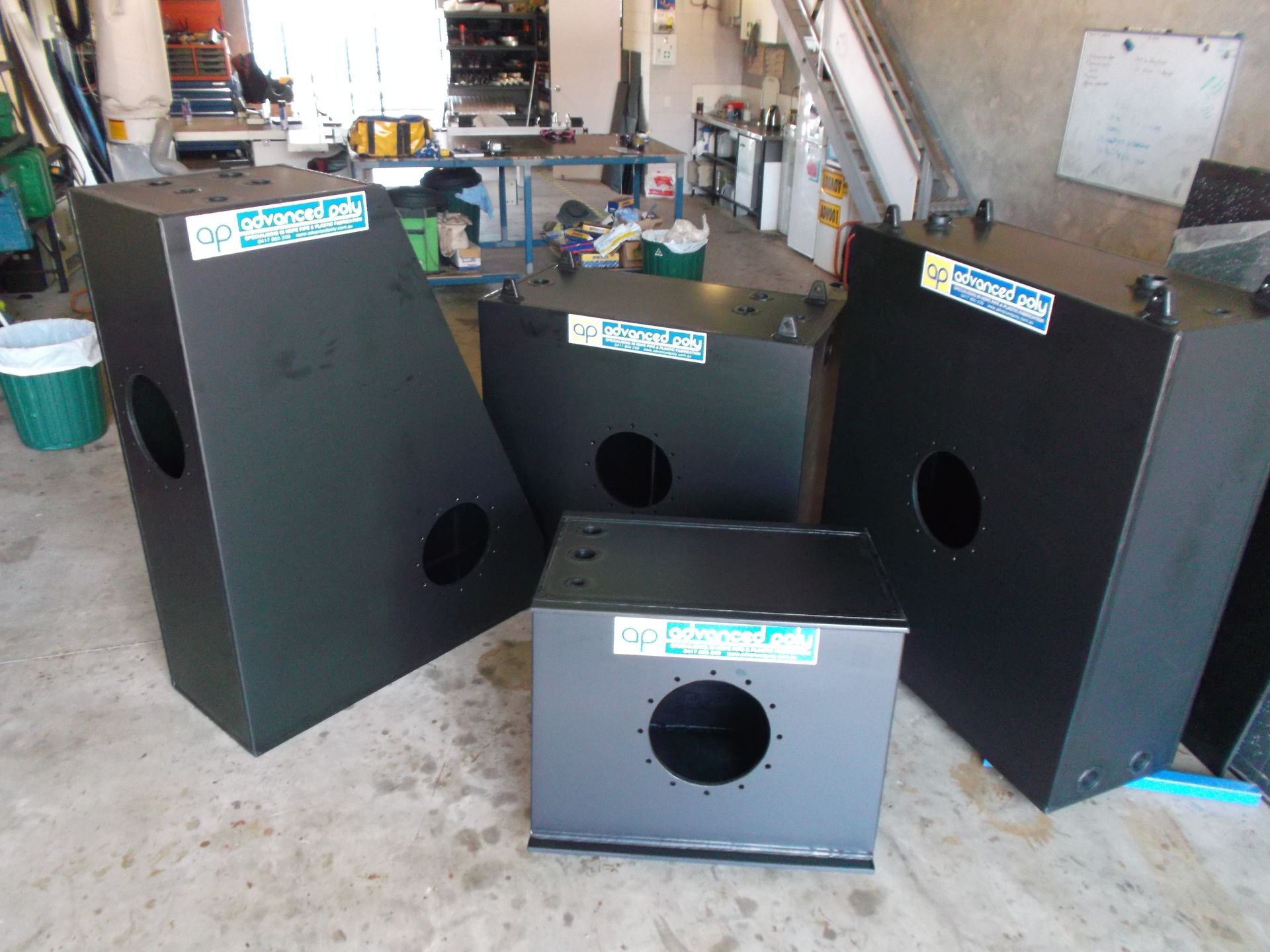 Four black metal speaker cabinets with circular cutouts arranged in a workshop garage. — Advanced Poly in Darwin, NT