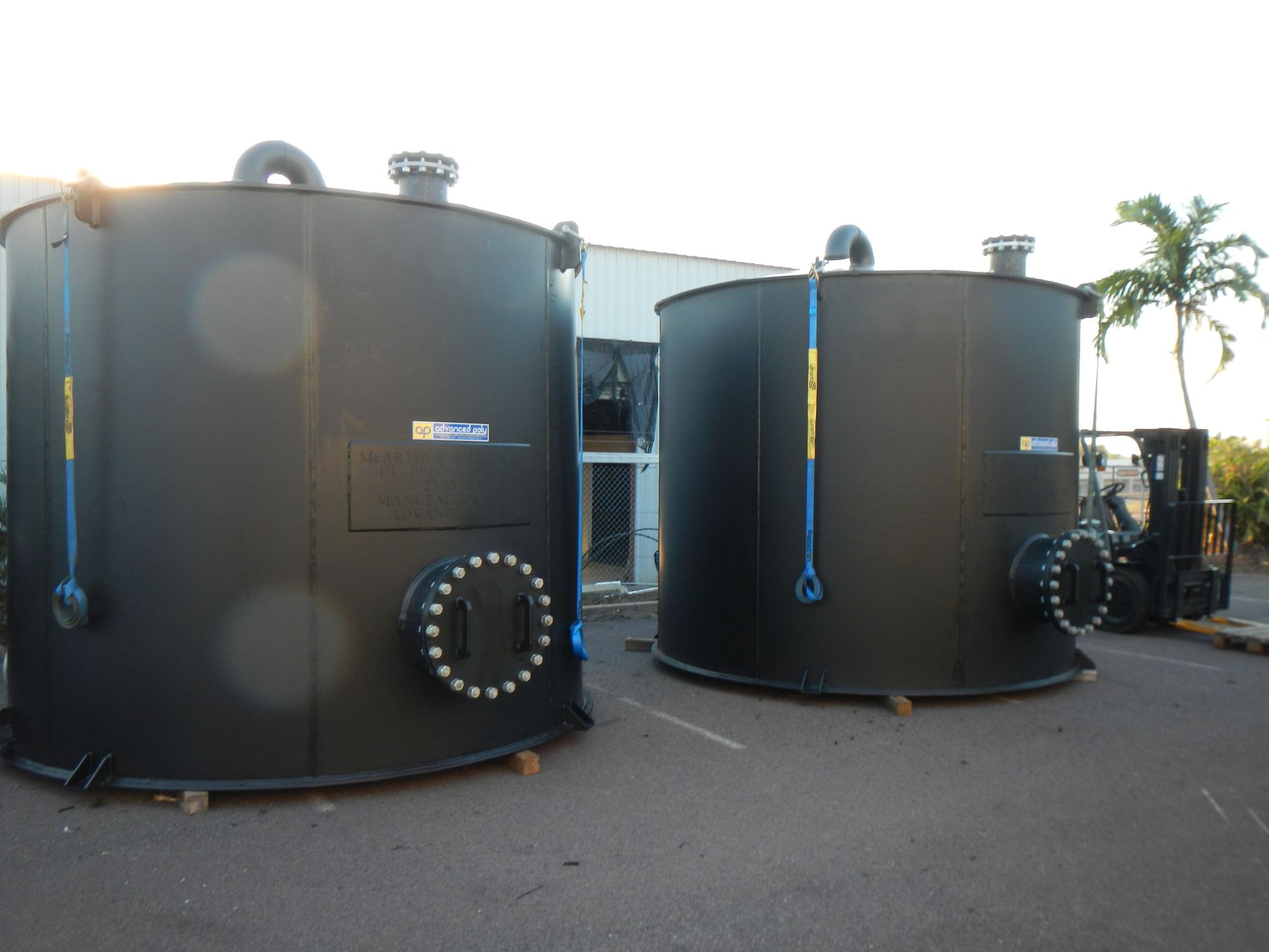 Two large black industrial tanks outdoors on a paved lot, with blue hoses and a forklift nearby. — Advanced Poly in Darwin, NT