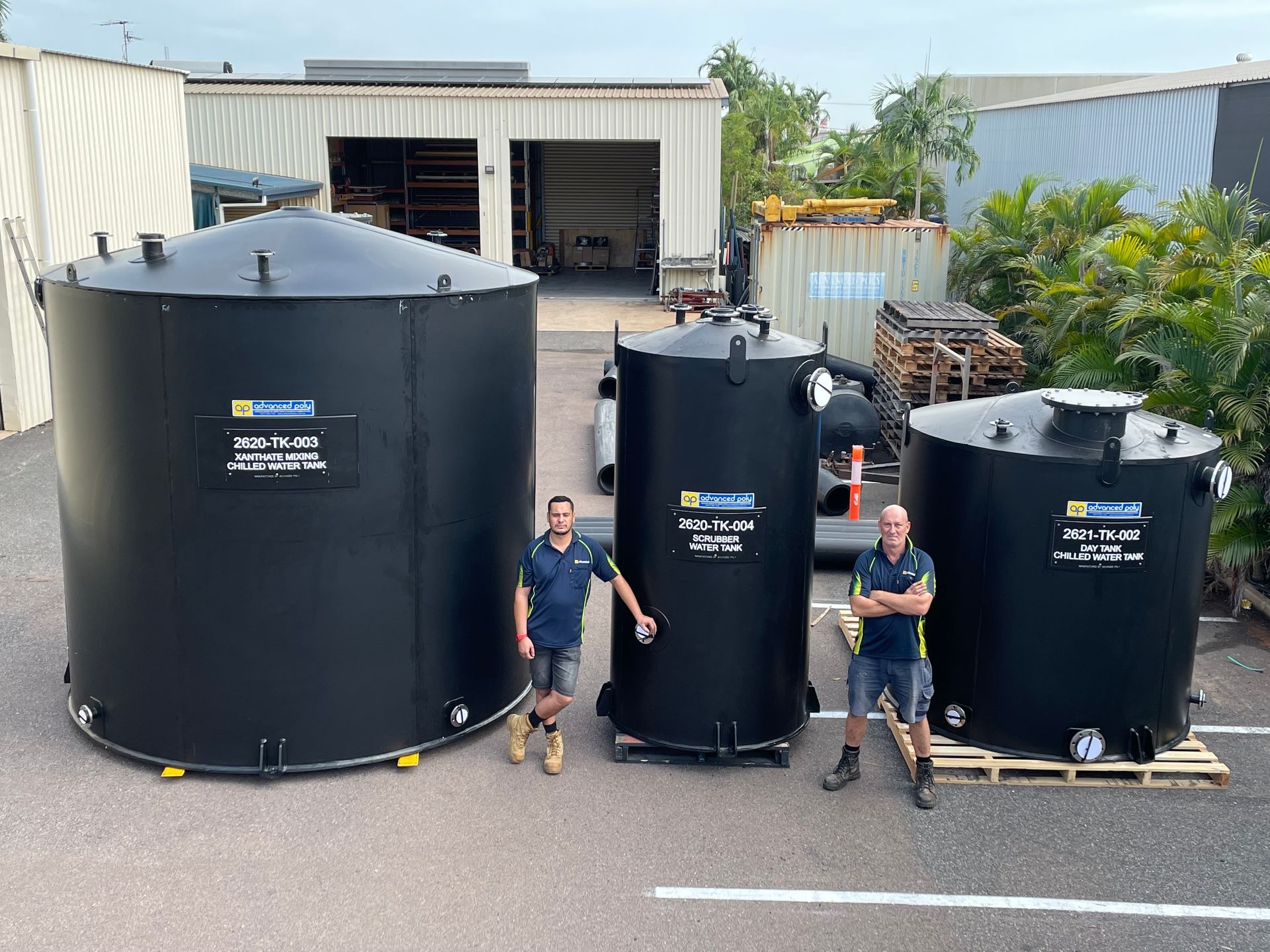 Three large black water tanks outdoors, with two people standing beside them in front of a warehouse. — Advanced Poly in Darwin, NT