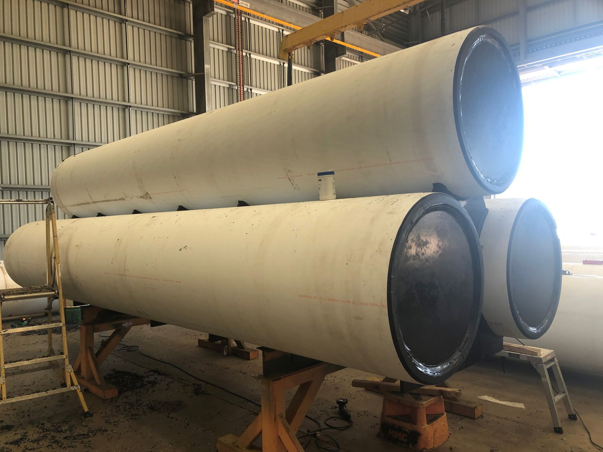 Two large white cylindrical tanks stacked in an industrial warehouse, with metal supports and shelving nearby — Advanced Poly in Darwin, NT