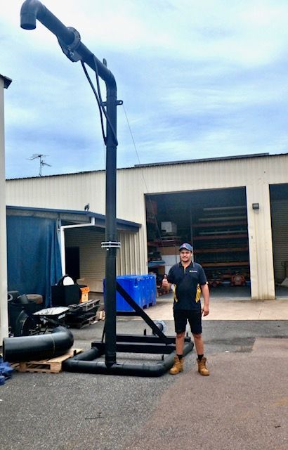 Person standing beside a tall black training rig in an industrial yard, wearing dark work clothes and boots. — Advanced Poly in Darwin, NT