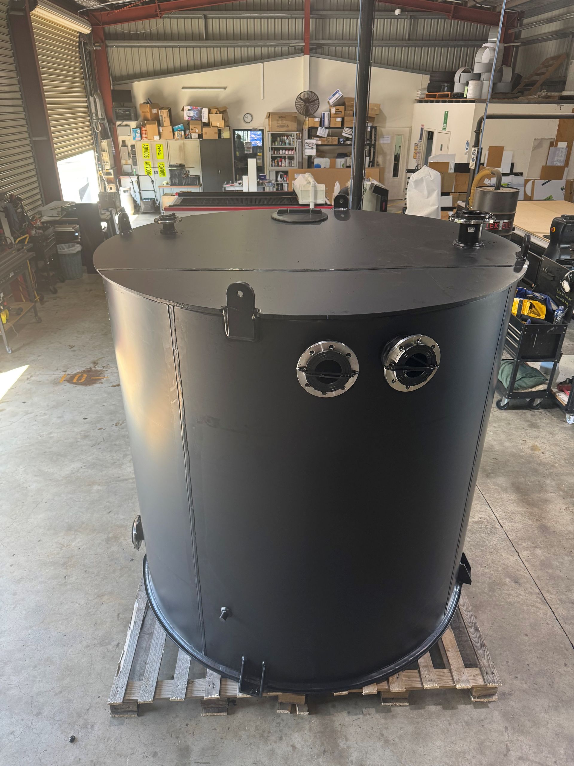 Large black cylindrical metal tank with two round ports, sitting on a pallet in a workshop. — Advanced Poly in Darwin, NT