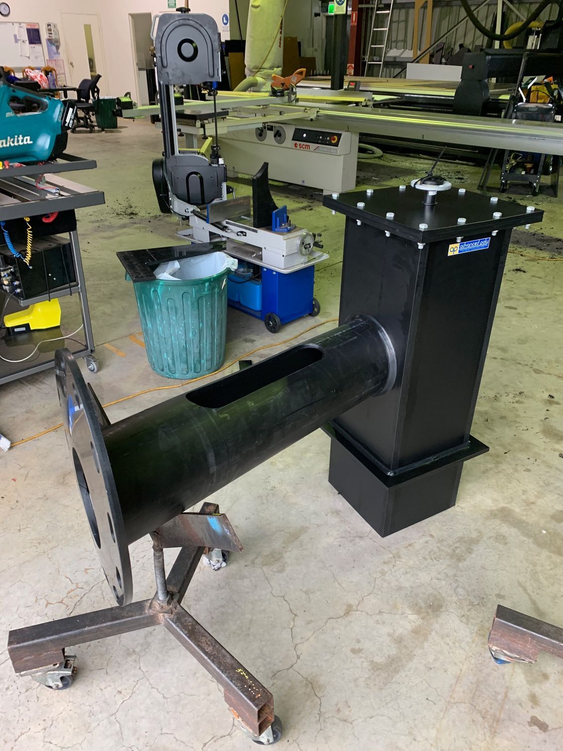 Black metal furnace on a stand in a workshop, with a long pipe extending left and tools/machinery in the background — Advanced Poly in Darwin, NT — Advanced Poly in Darwin, NT — Advanced Poly in Darwin, NT — Advanced Poly in Darwin, NT — Advanced Poly in Darwin, NT 