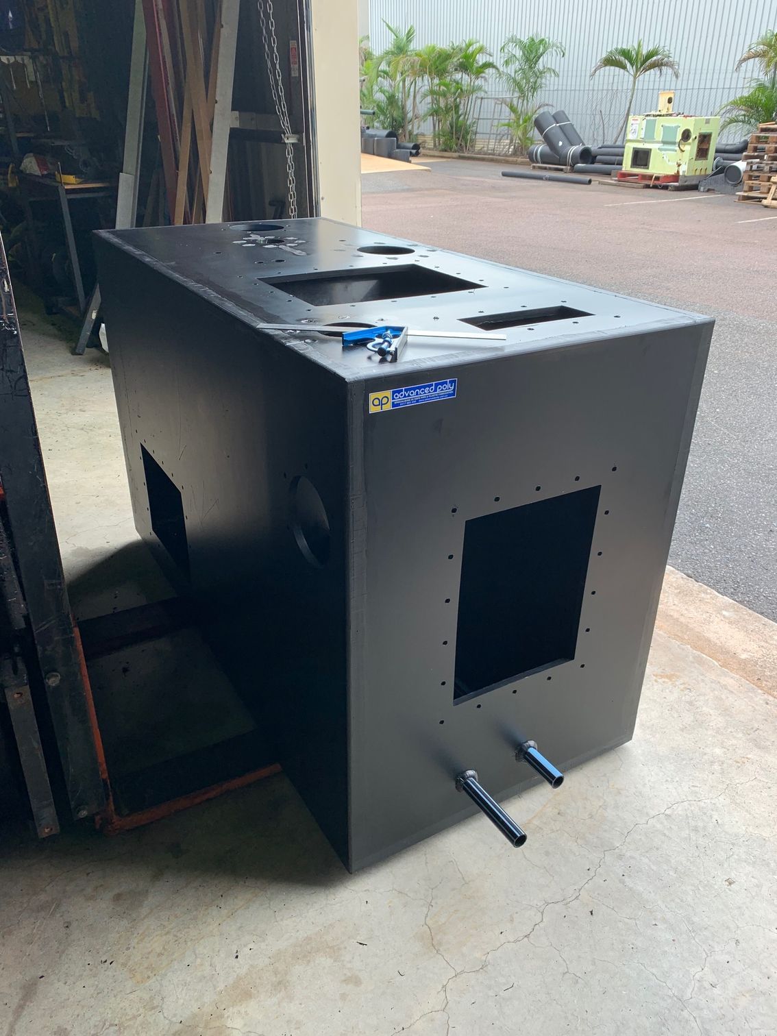Black metal industrial machine or cabinet outdoors beside a truck, with cutout openings and blue label — Advanced Poly in Darwin, NT — Advanced Poly in Darwin, NT — Advanced Poly in Darwin, NT — Advanced Poly in Darwin, NT — Advanced Poly in Darwin, NT 