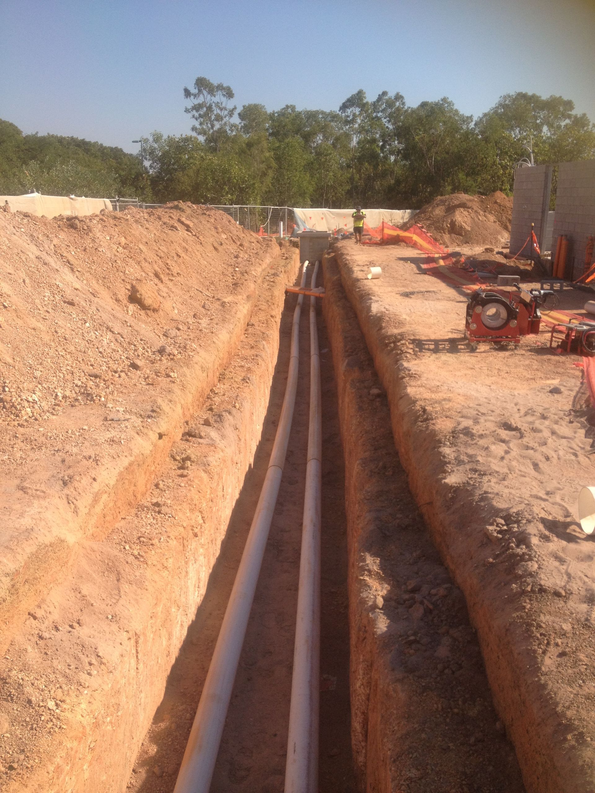 Trenched construction site with long narrow ditch, dirt piles, and heavy equipment under a clear sky — Advanced Poly in Darwin, NT