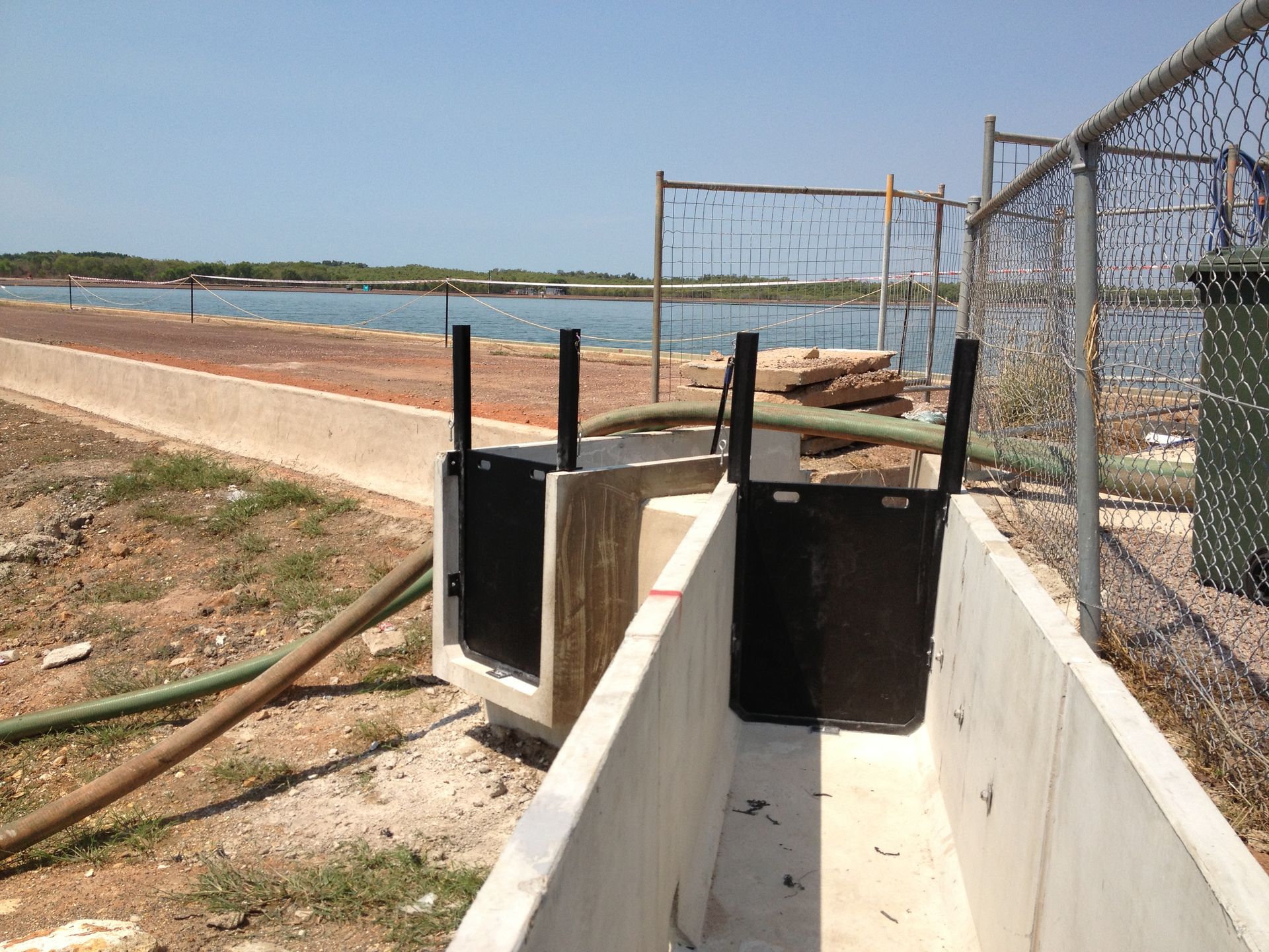 Concrete drainage channel beside a waterfront path and chain-link fence under clear sky — Advanced Poly in Darwin, NT