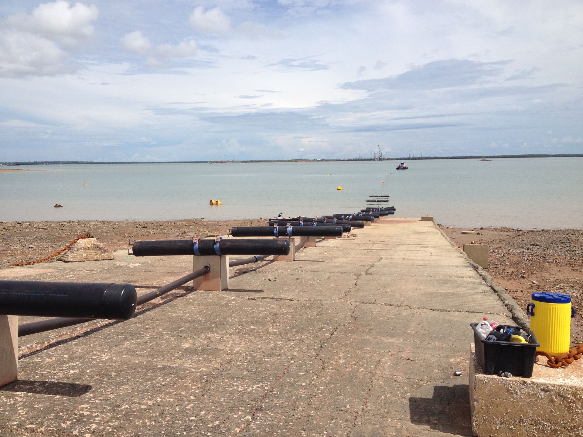 Concrete pier with black pipes leading into calm sea, sandy beach, and cloudy sky — Advanced Poly in Darwin, NT