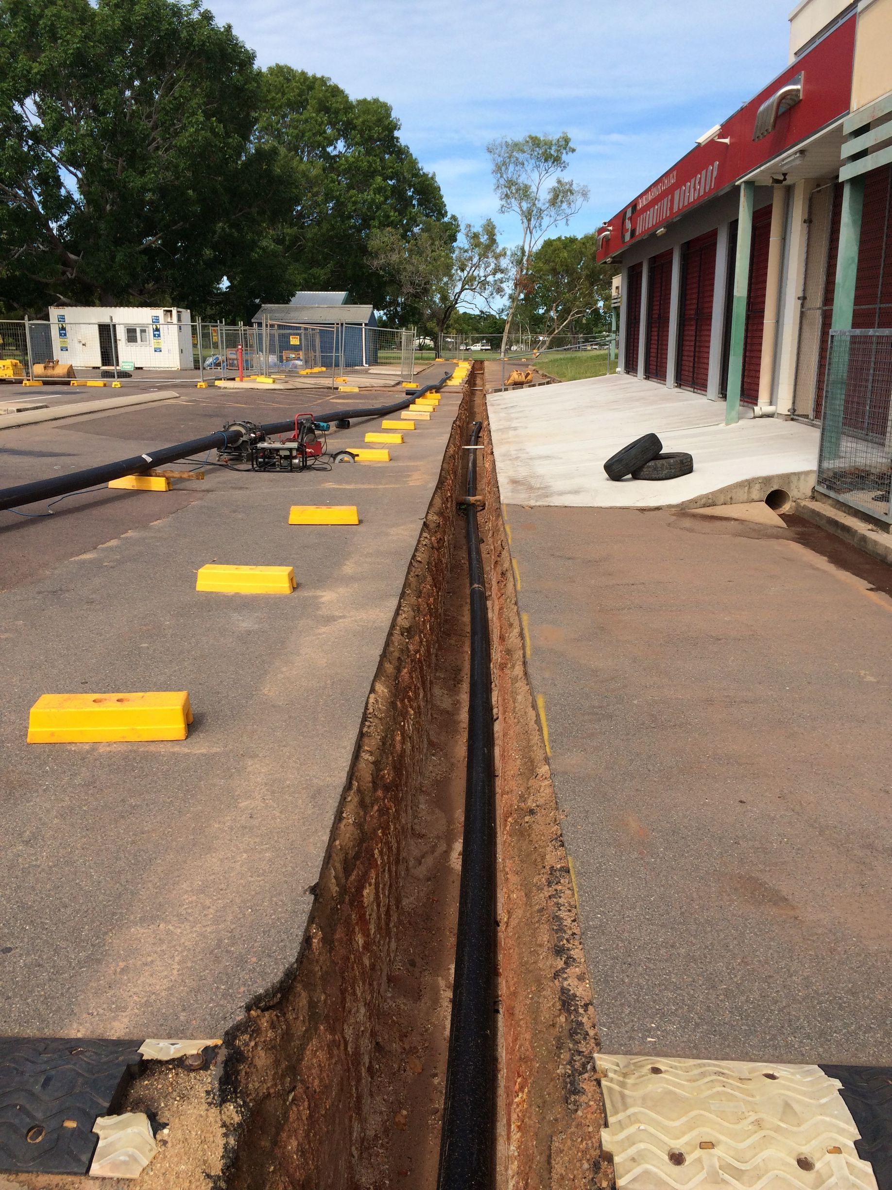 Cracked parking lot beside storefronts with a long open trench and yellow markers, suggesting road repair — Advanced Poly in Darwin, NT