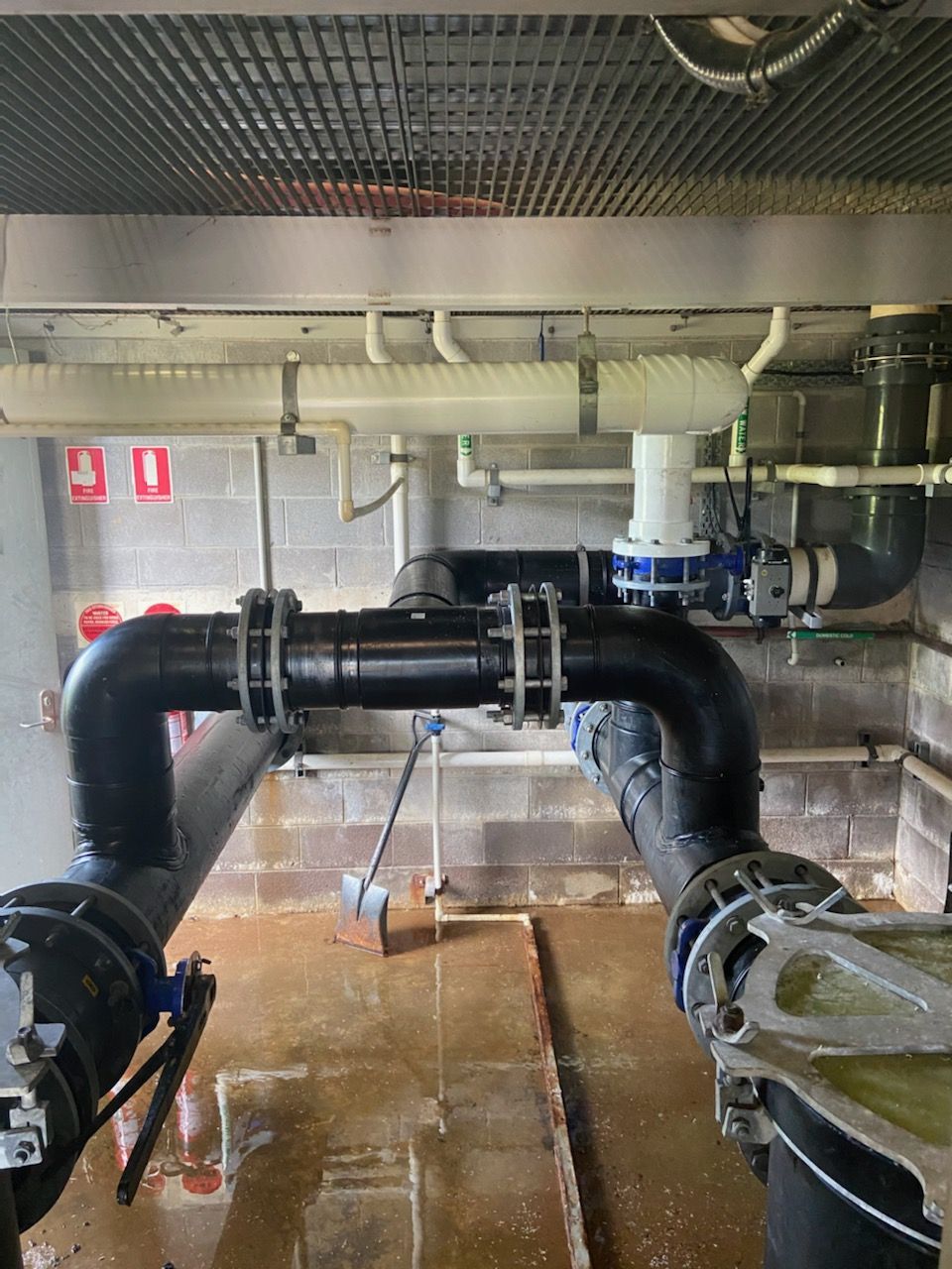 Industrial pipework in a utility room, with large black pipes, valves, and a wet floor against a concrete wall. — Advanced Poly in Darwin, NT
