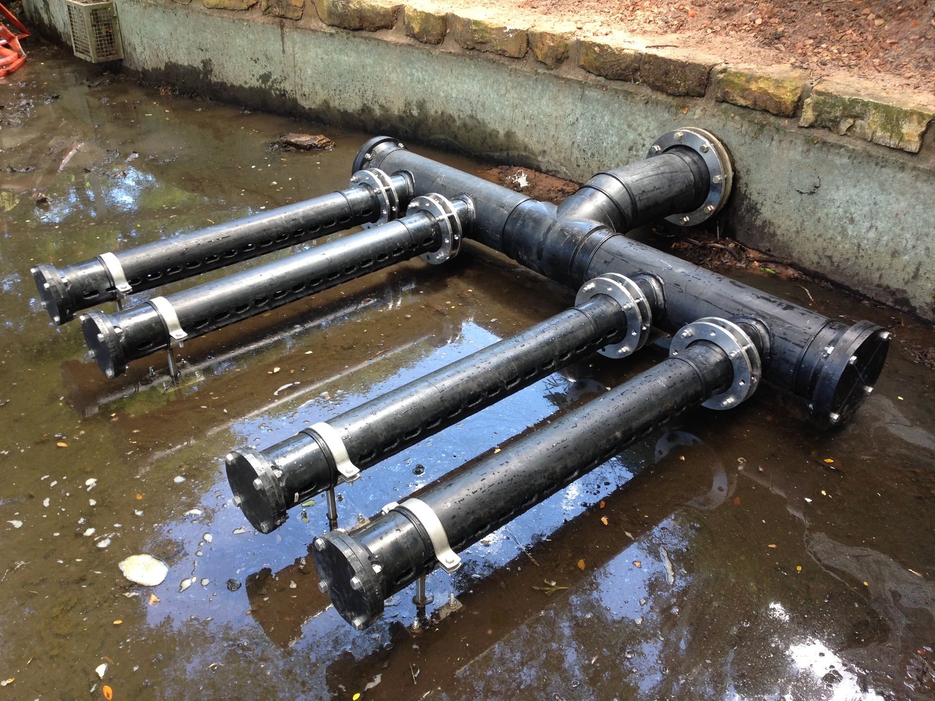 Three black metal pipes lying in shallow water beside a brick wall — Advanced Poly in Darwin, NT