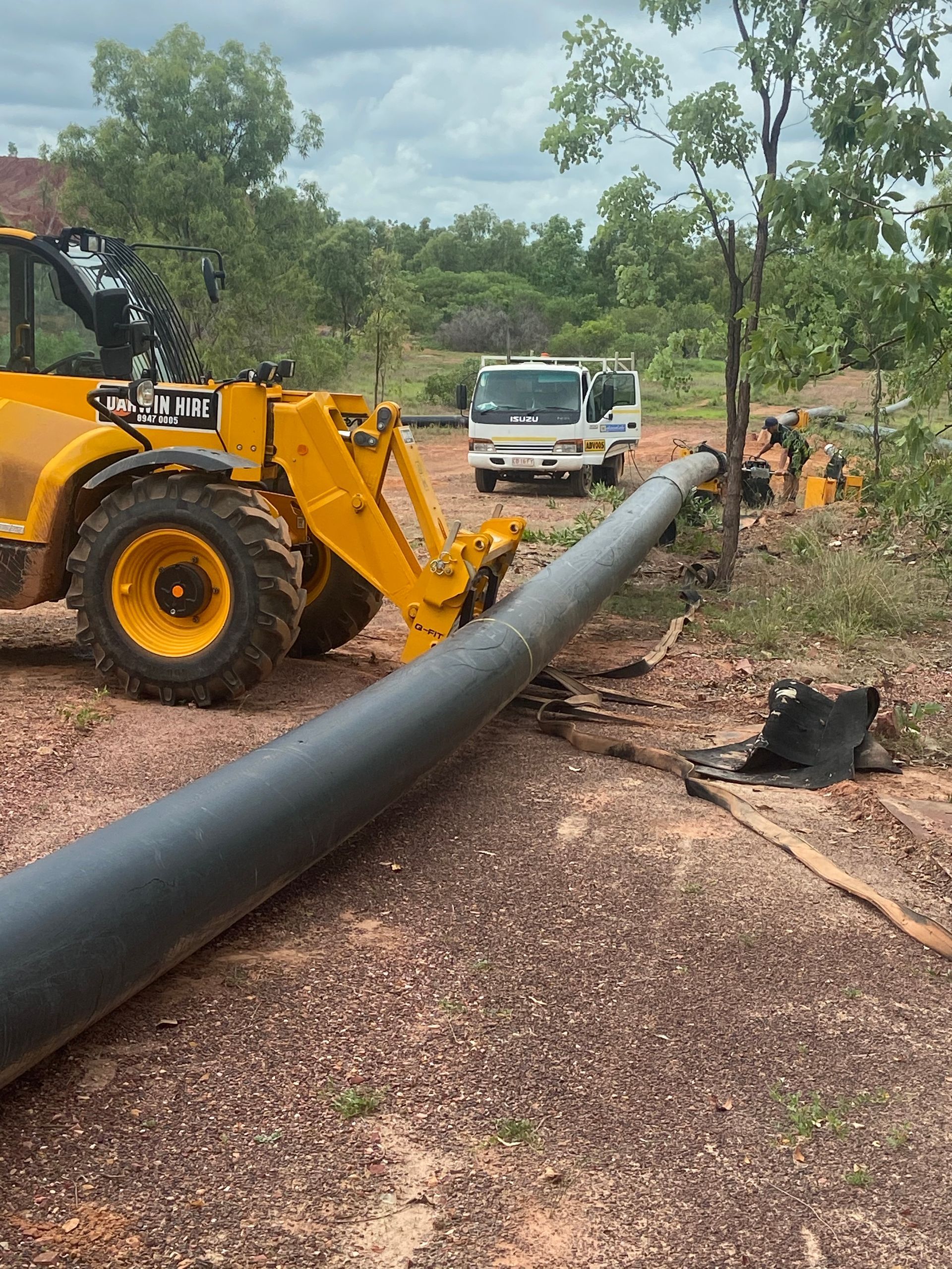 Yellow construction loader moving a large black pipe on a muddy work site with a truck and trees nearby — Advanced Poly in Darwin, NT