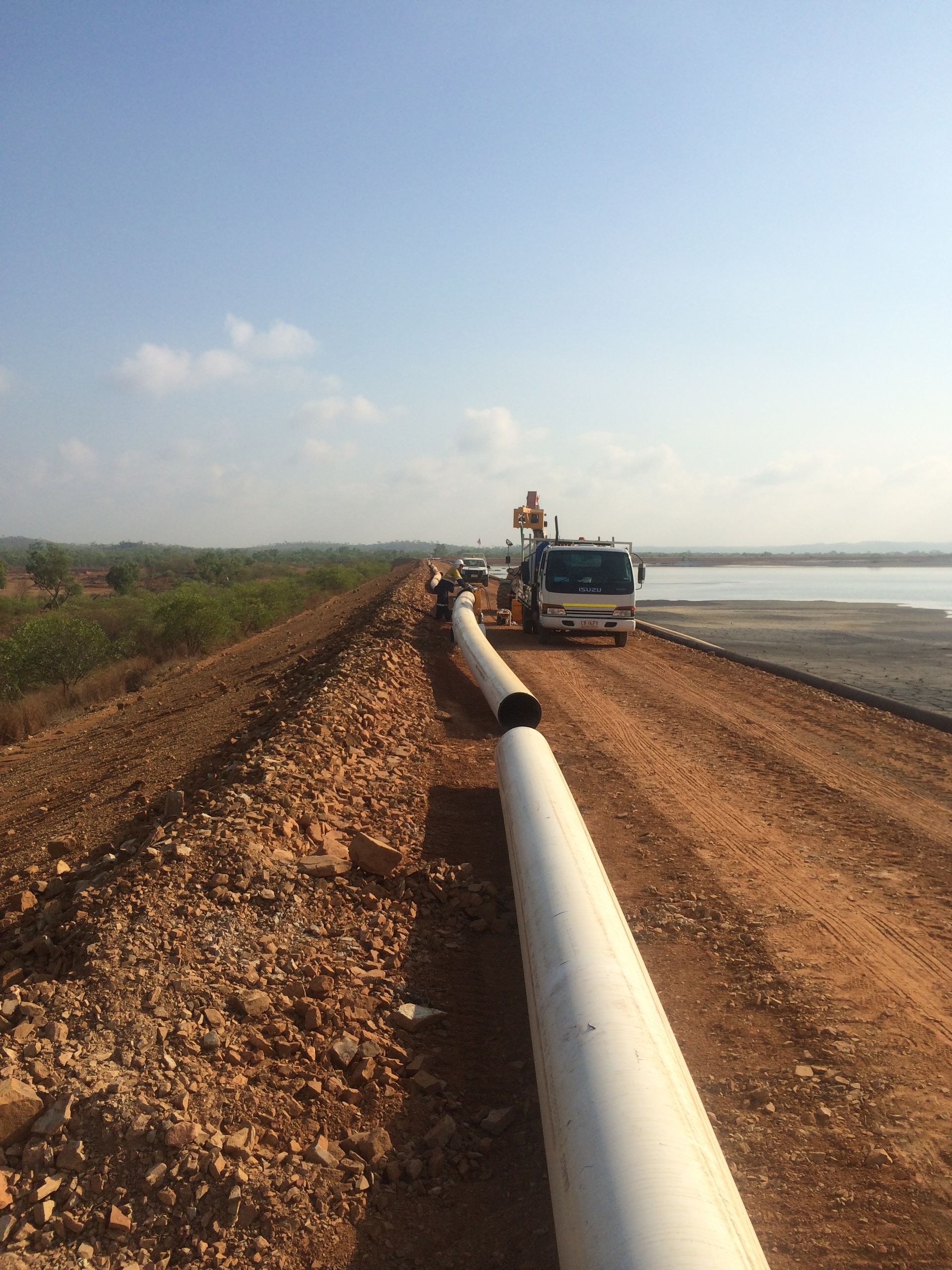 Pipeline construction along a dirt road beside water, with heavy machinery in the distance. — Advanced Poly in Darwin, NT