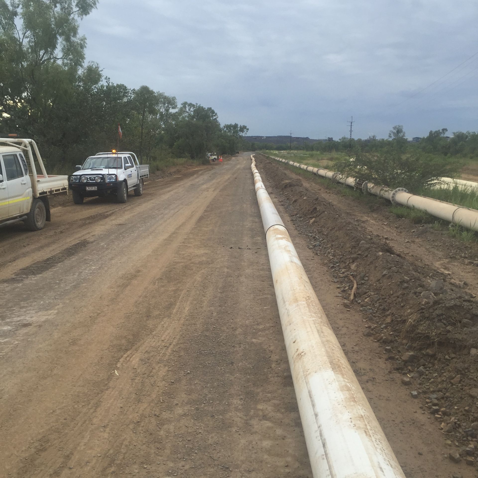 Dirt road beside a large white pipeline, with parked pickup trucks and cloudy sky overhead. — Advanced Poly in Darwin, NT