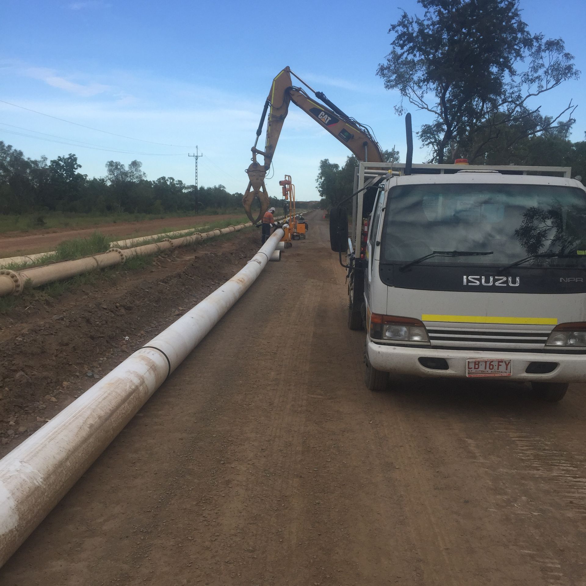 Roadwork site with an Isuzu truck, excavator, and long white pipeline on a dirt road under construction — Advanced Poly in Darwin, NT