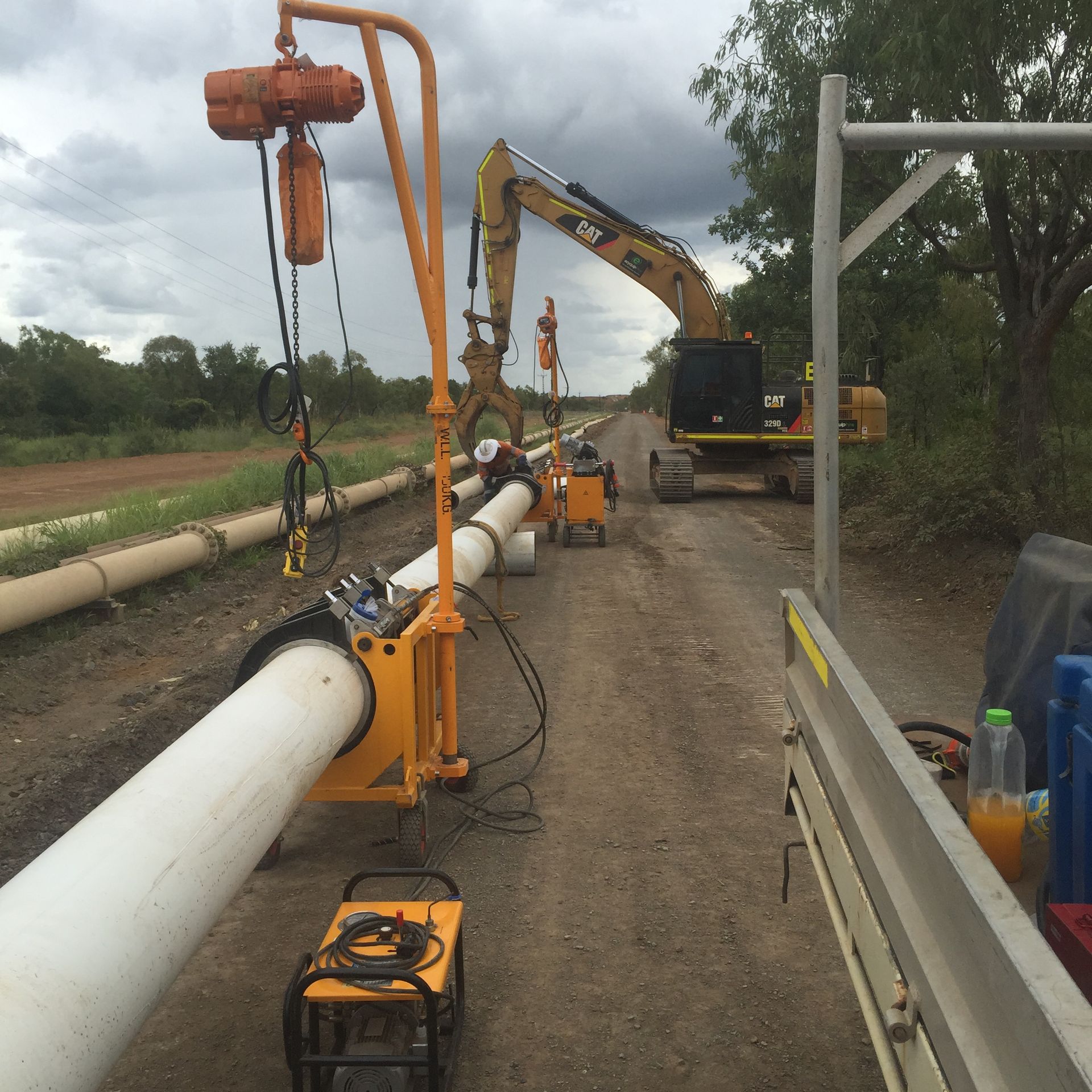 Pipeline construction site with white pipe sections, orange lifting rigs, excavator, and workers along a dirt road — Advanced Poly in Darwin, NT