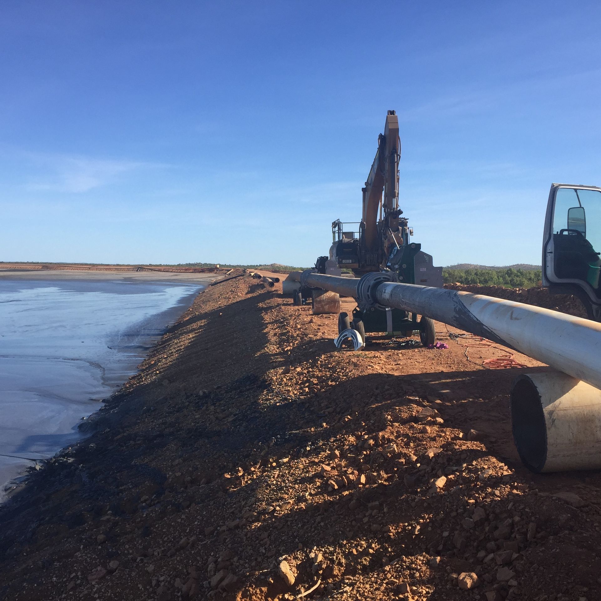 Pipeline and excavator on a gravel embankment beside a calm body of water under a clear blue sky — Advanced Poly in Darwin, NT