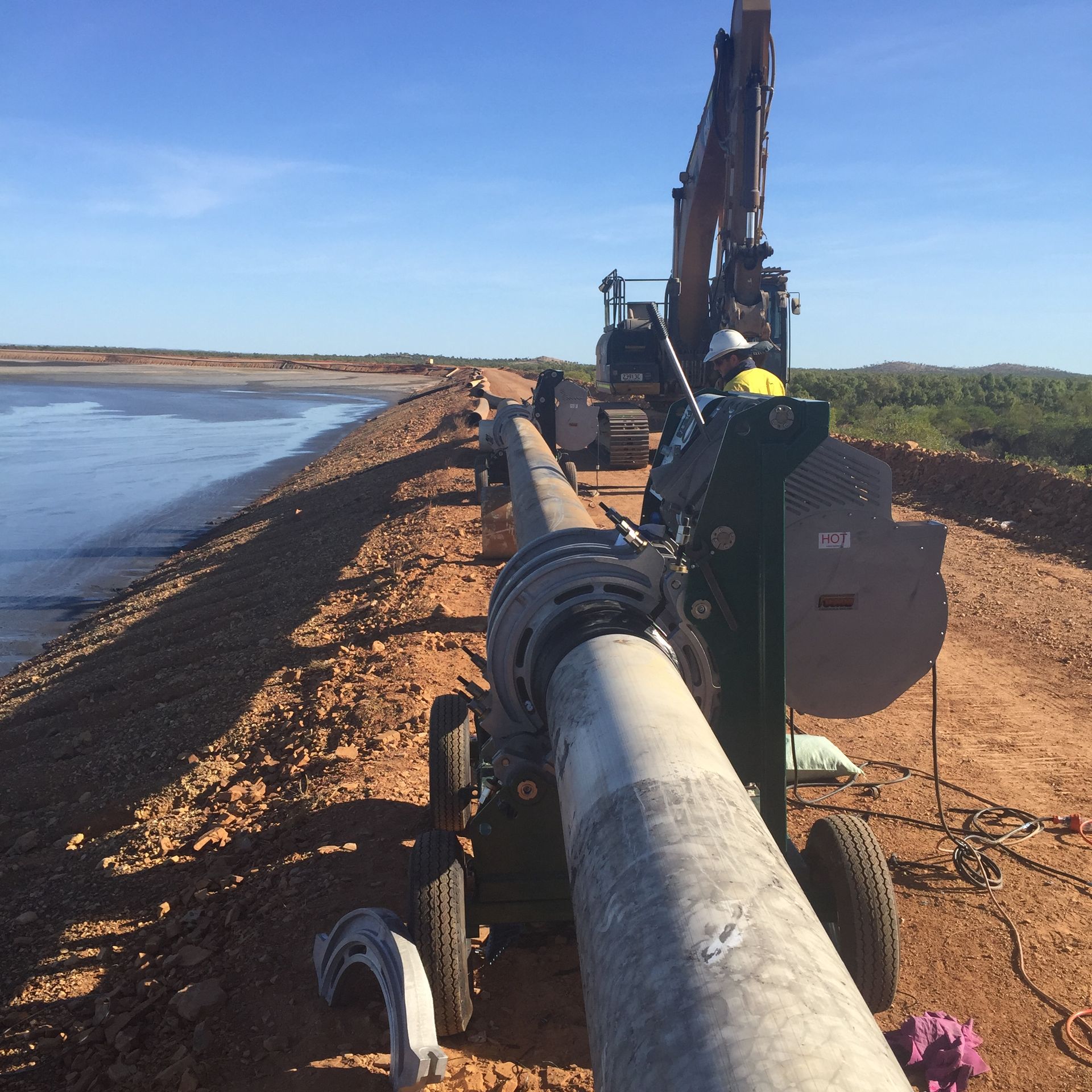 Heavy machinery laying a large pipe along a dirt embankment beside water under a clear blue sky — Advanced Poly in Darwin, NT