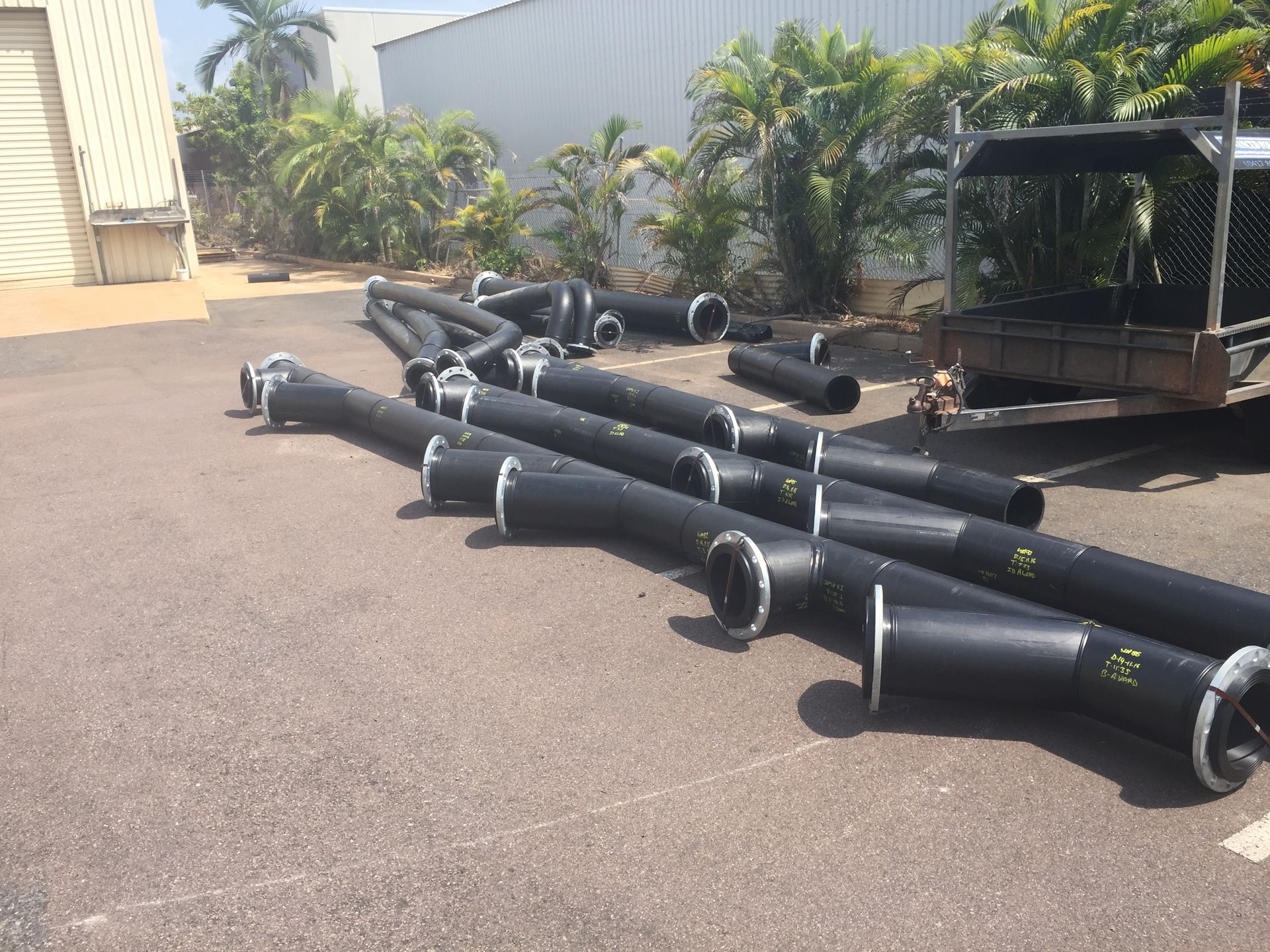 Several black metal pipes and fittings scattered on pavement beside a building and greenery — Advanced Poly in Darwin, NT