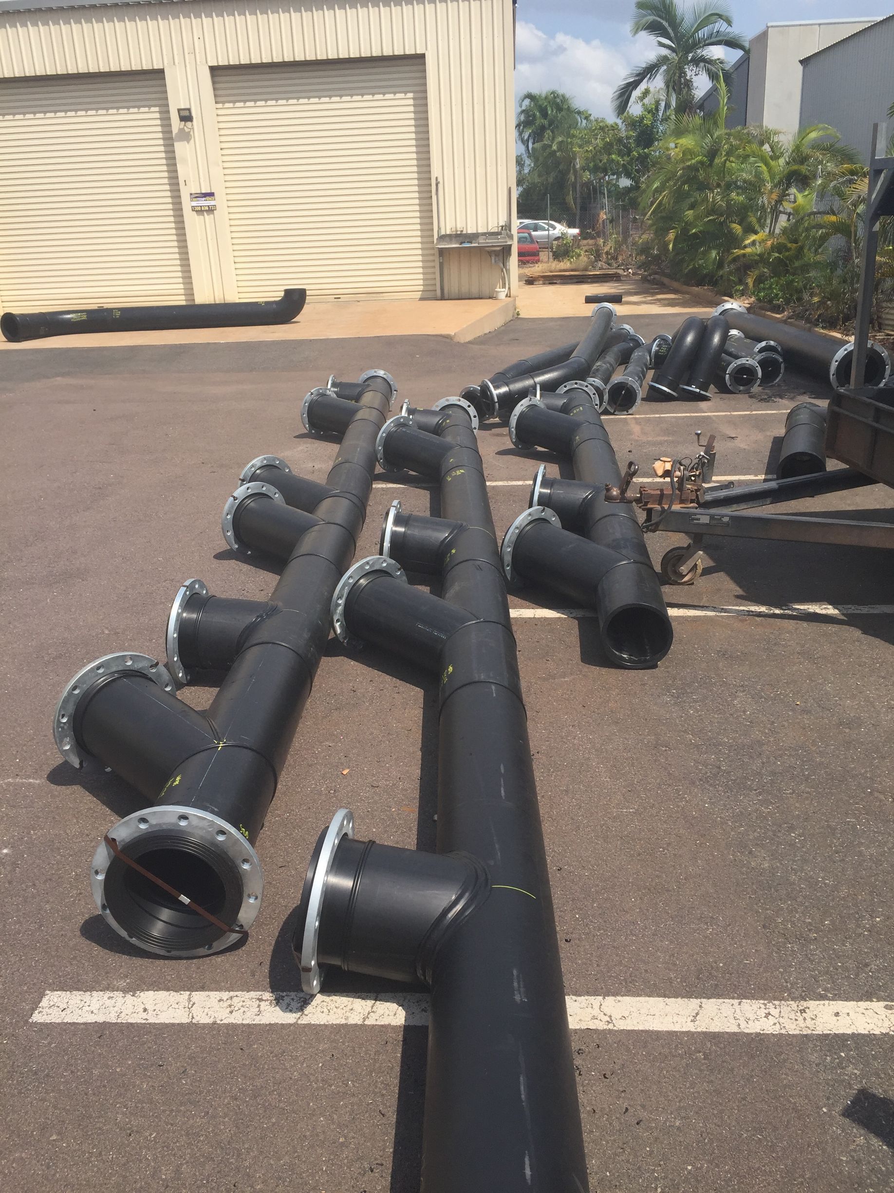 Black industrial pipes with flanges stacked on asphalt near a beige warehouse on a sunny day — Advanced Poly in Darwin, NT
