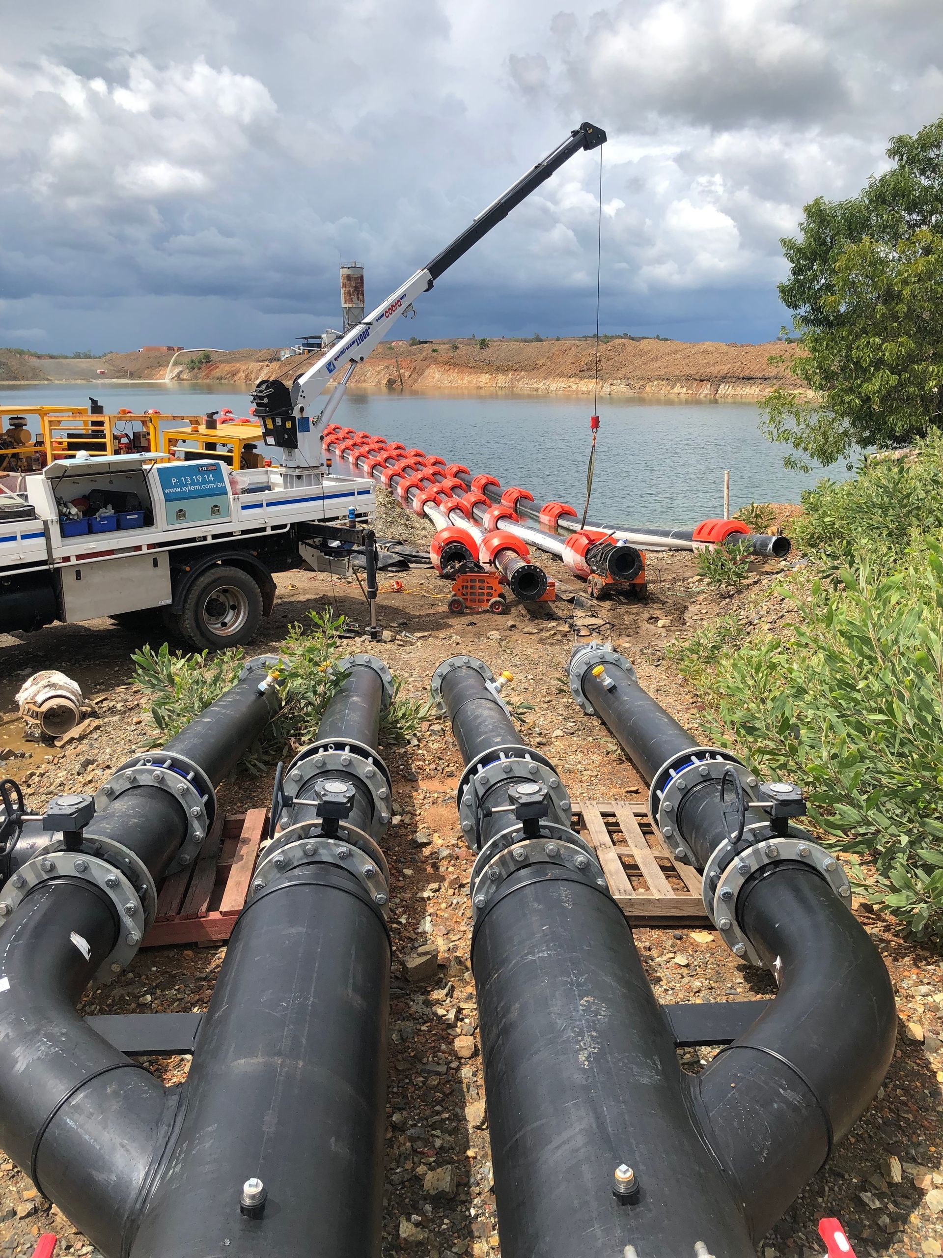 Large black pipelines and red-orange floating boom extending across a river from a work truck with crane — Advanced Poly in Darwin, NT