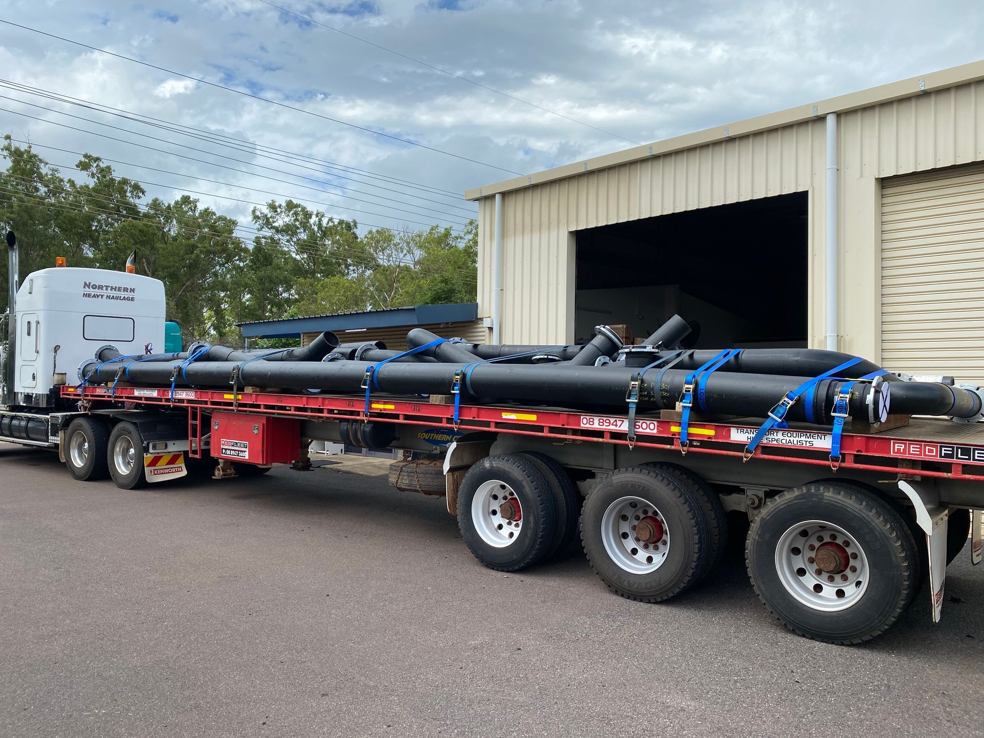 Long black pipe load strapped on a red flatbed trailer outside a warehouse — Advanced Poly in Darwin, NT 