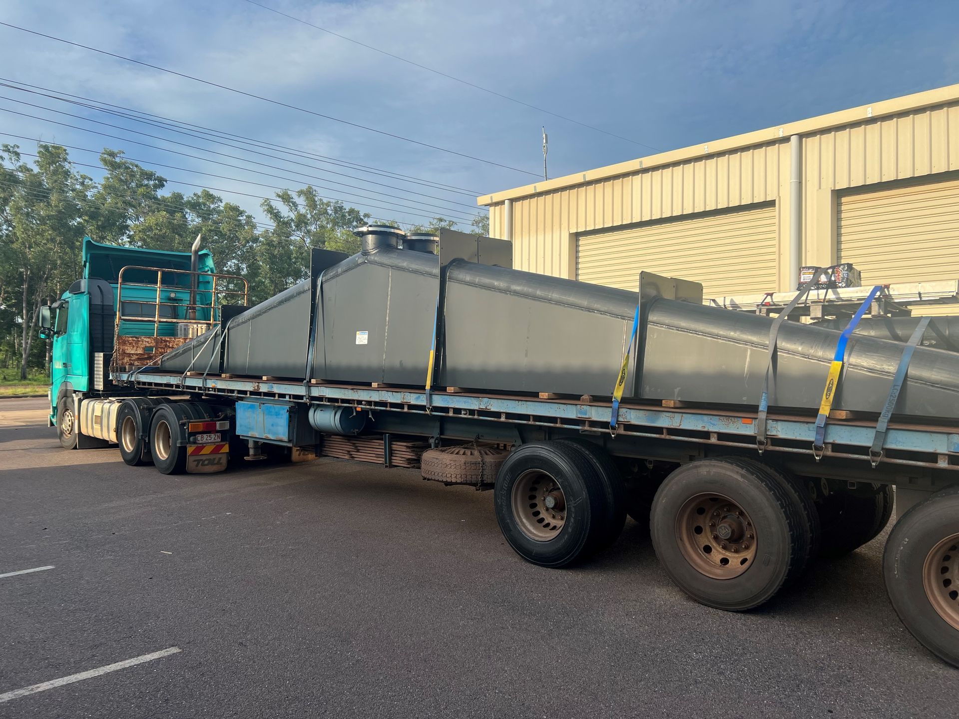 Flatbed truck carrying a long gray metal beam parked beside a yellow warehouse — Advanced Poly in Darwin, NT 