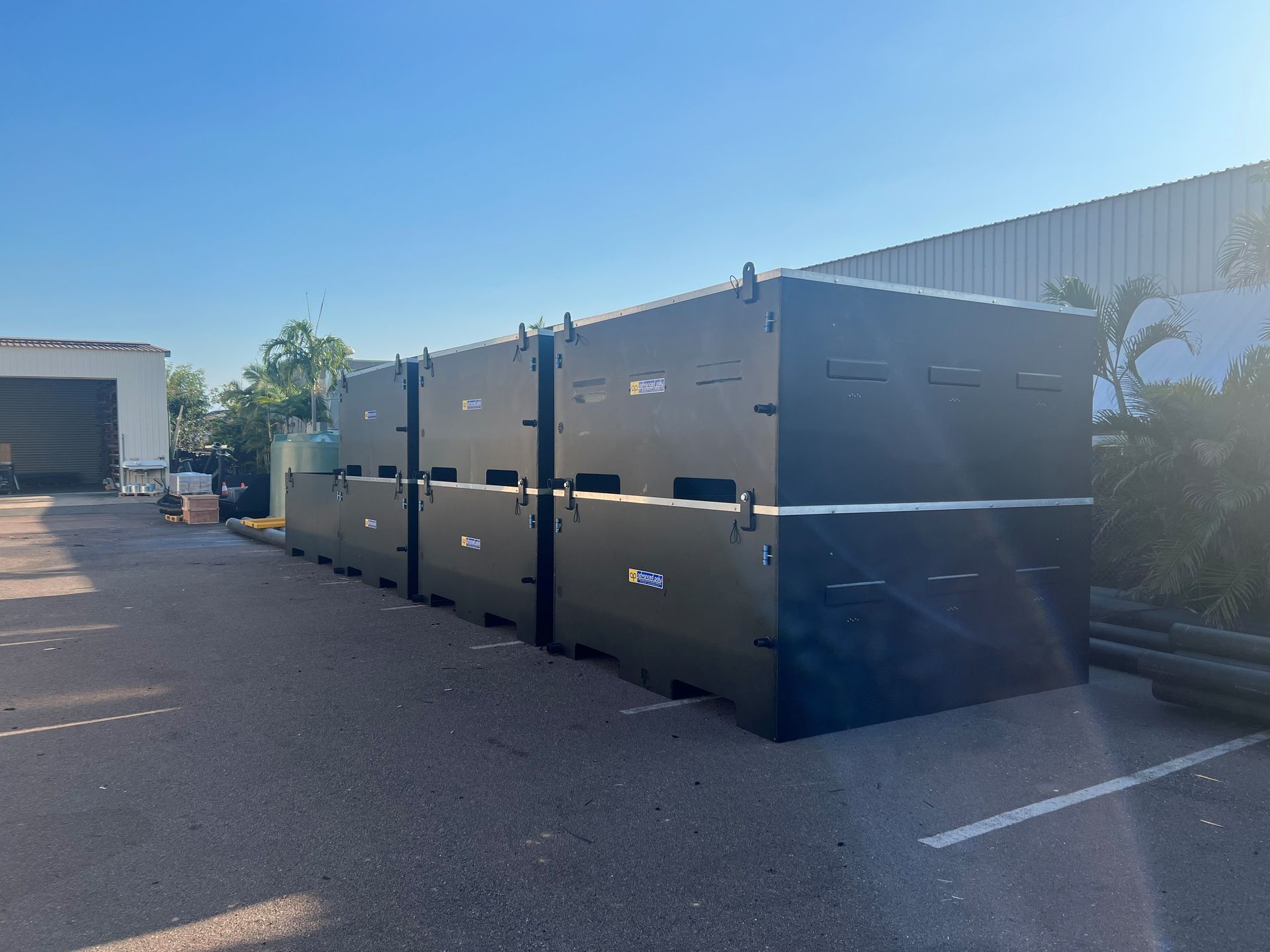 Row of large black industrial dumpsters beside a warehouse in a sunny parking lot — Advanced Poly in Darwin, NT