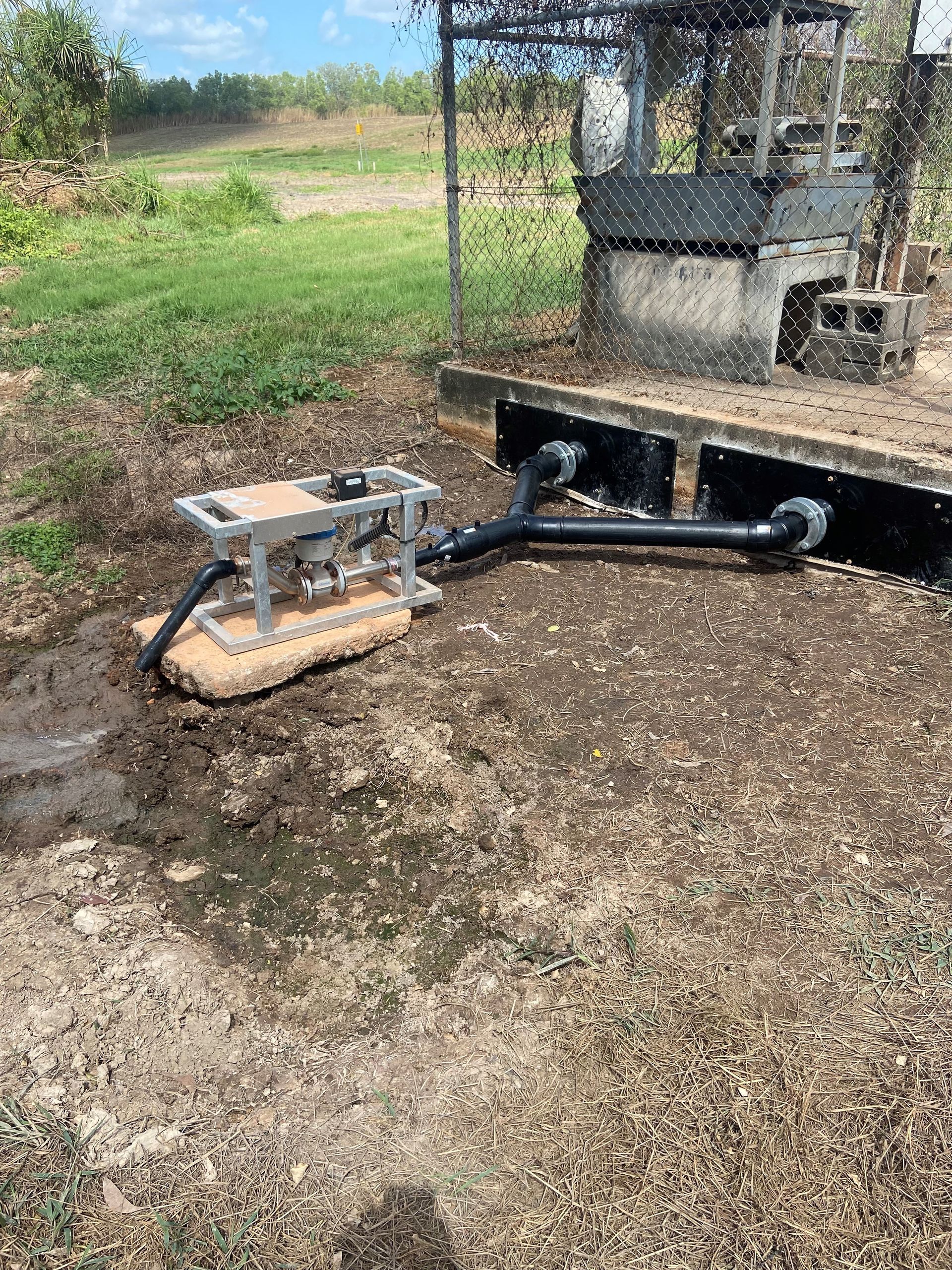 Black hose and metal pump mounted on a concrete base in a muddy outdoor field. — Advanced Poly in Darwin, NT