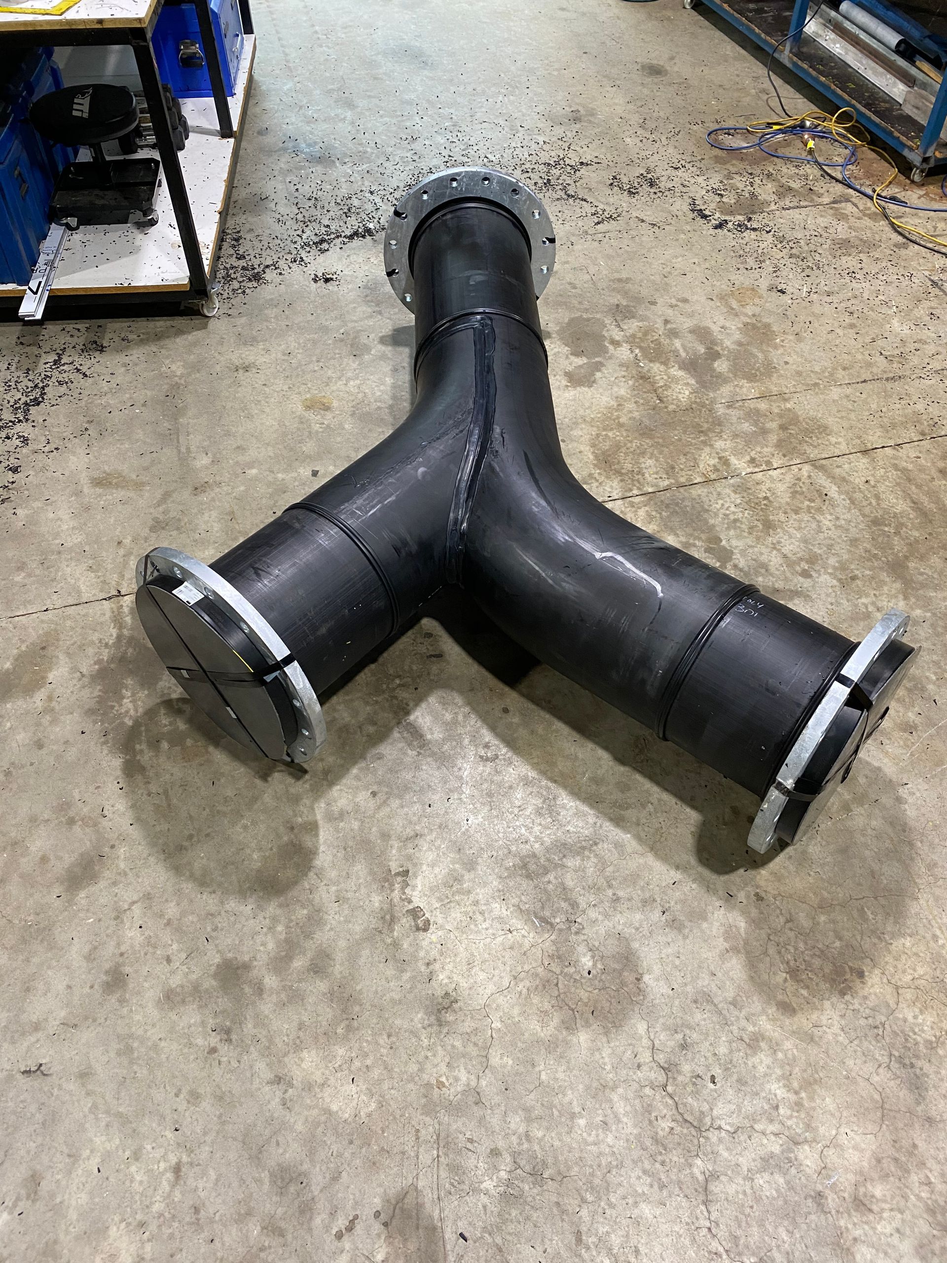 Black three-way pipe fitting on a concrete workshop floor, with metal flanges at each end. — Advanced Poly in Darwin, NT