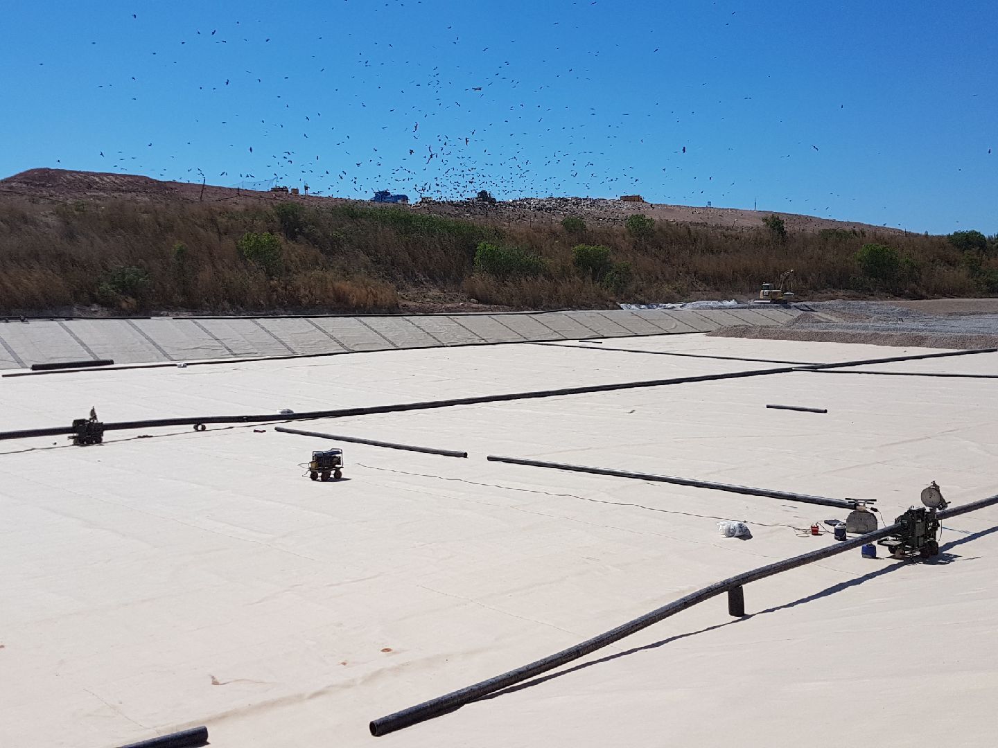 White rooftop with seams and scattered equipment under a clear blue sky, with a tree-lined hillside in back — Advanced Poly in Darwin, NT