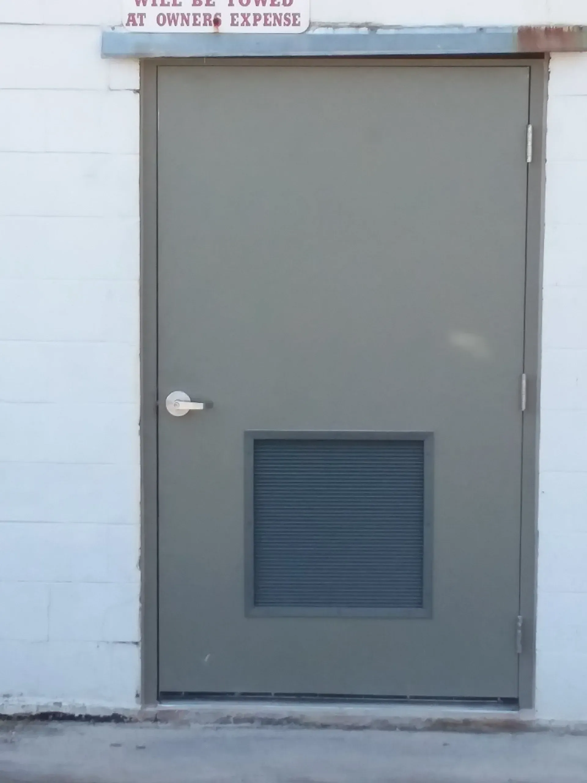 Gray metal door with a small vent panel in a white wall.