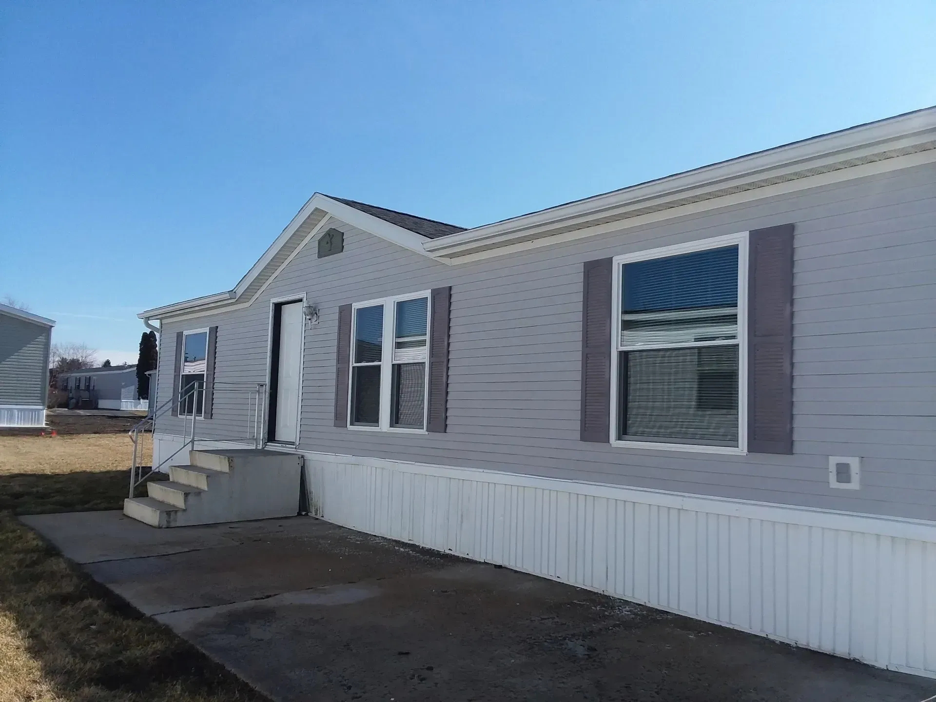 White manufactured home with small front steps, gray shutters, and a paved driveway under a clear sky