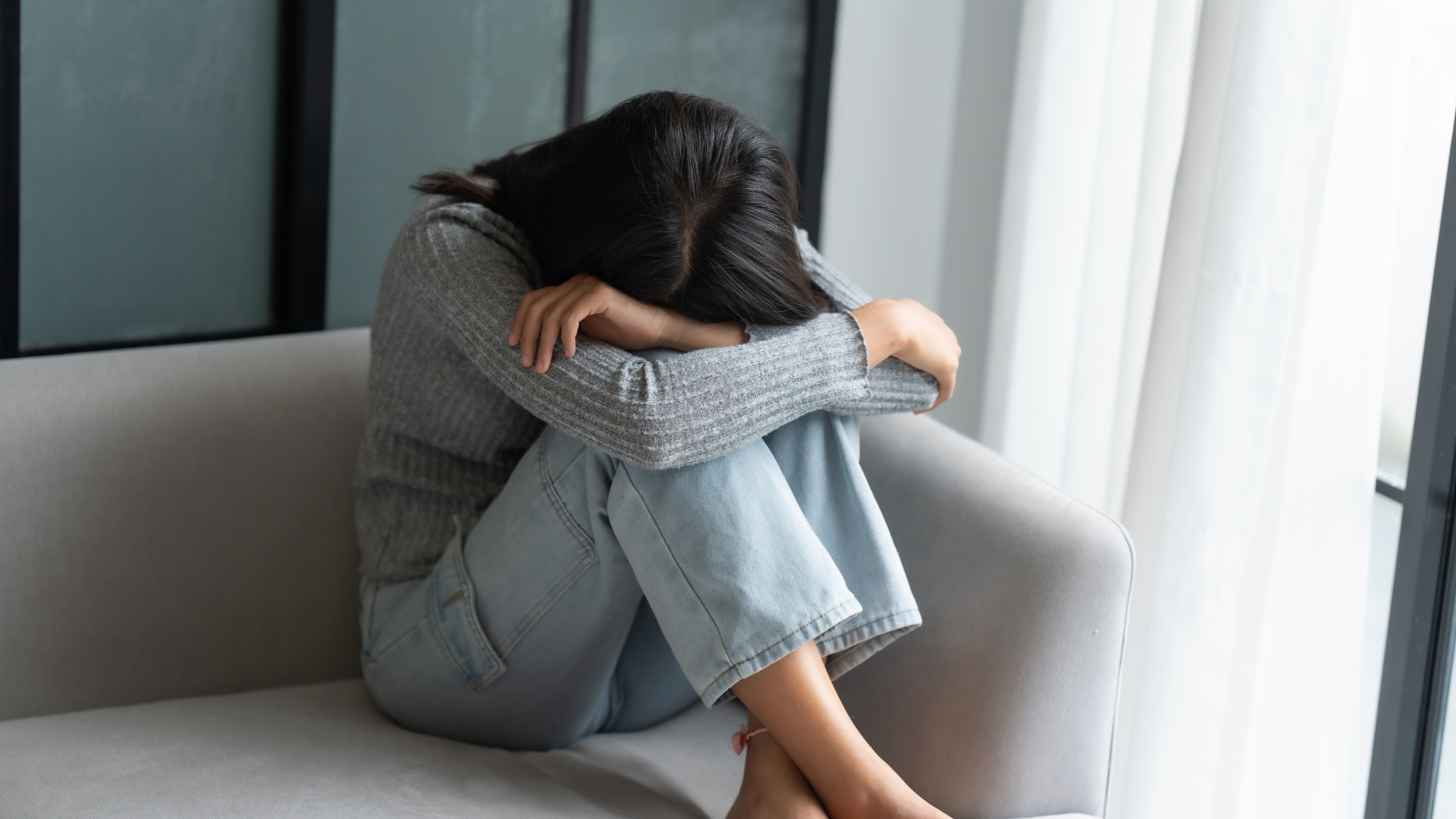 A person sitting on a sofa with their head tucked into their knees, arms wrapped around their legs.