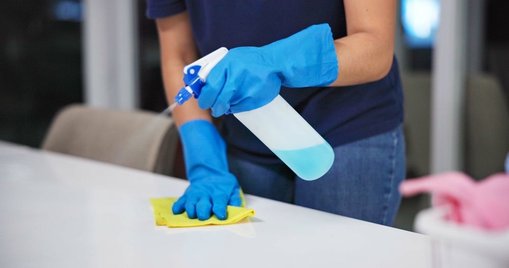 Person in blue gloves sprays cleaner and wipes a white table with a yellow cloth.