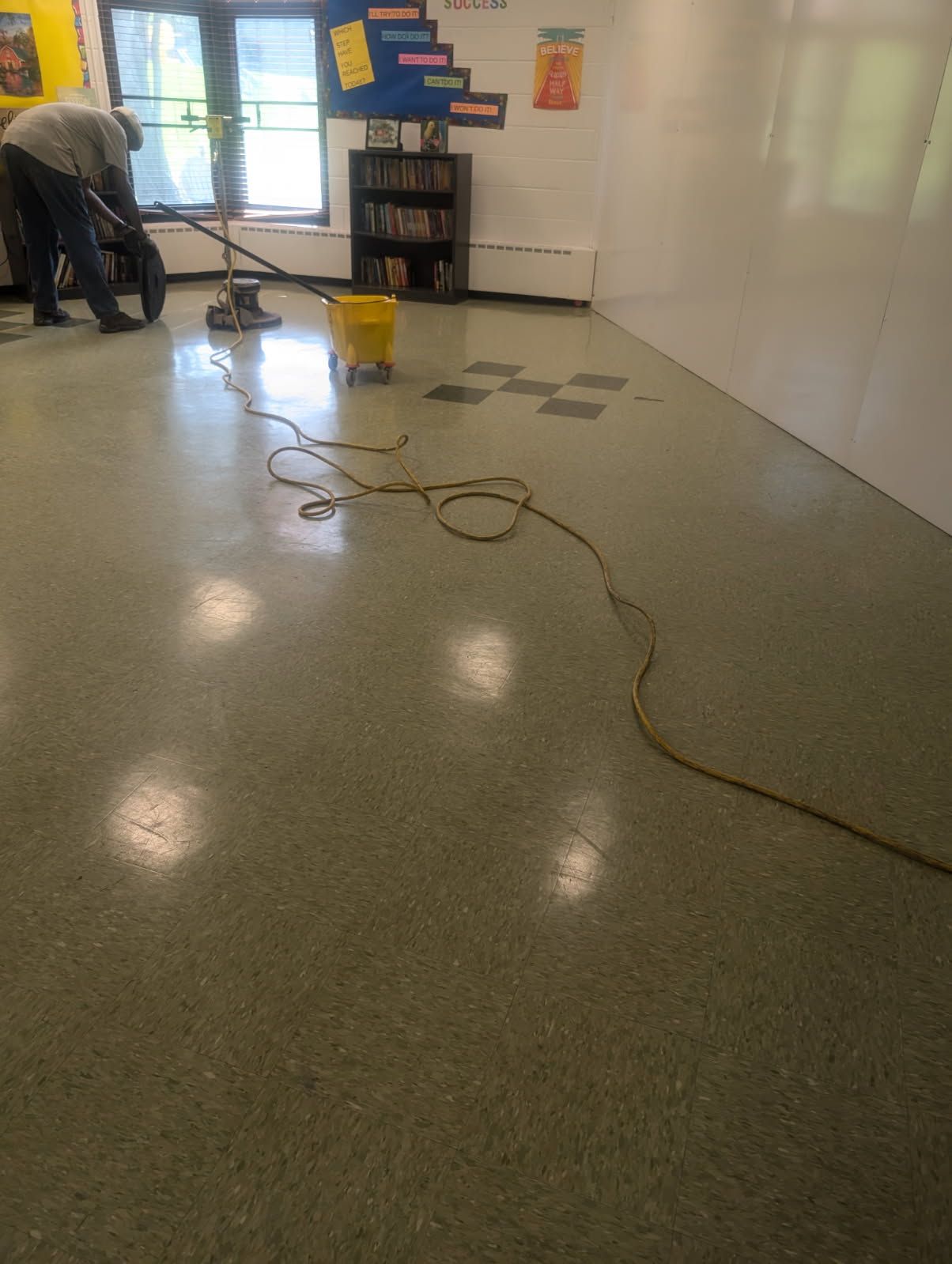 Person cleaning a glossy classroom floor with a mop and bucket near windows and shelves