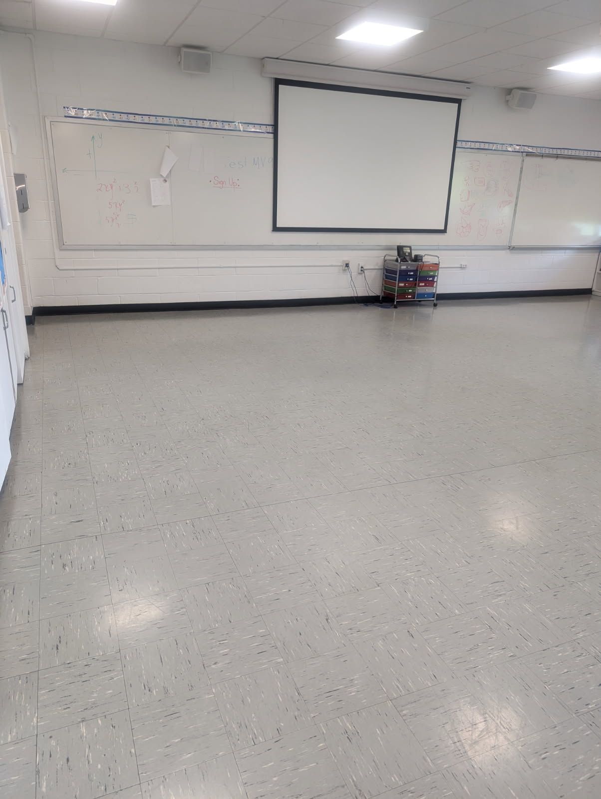 Empty classroom with whiteboard, pull-down projector screen, and gray speckled floor